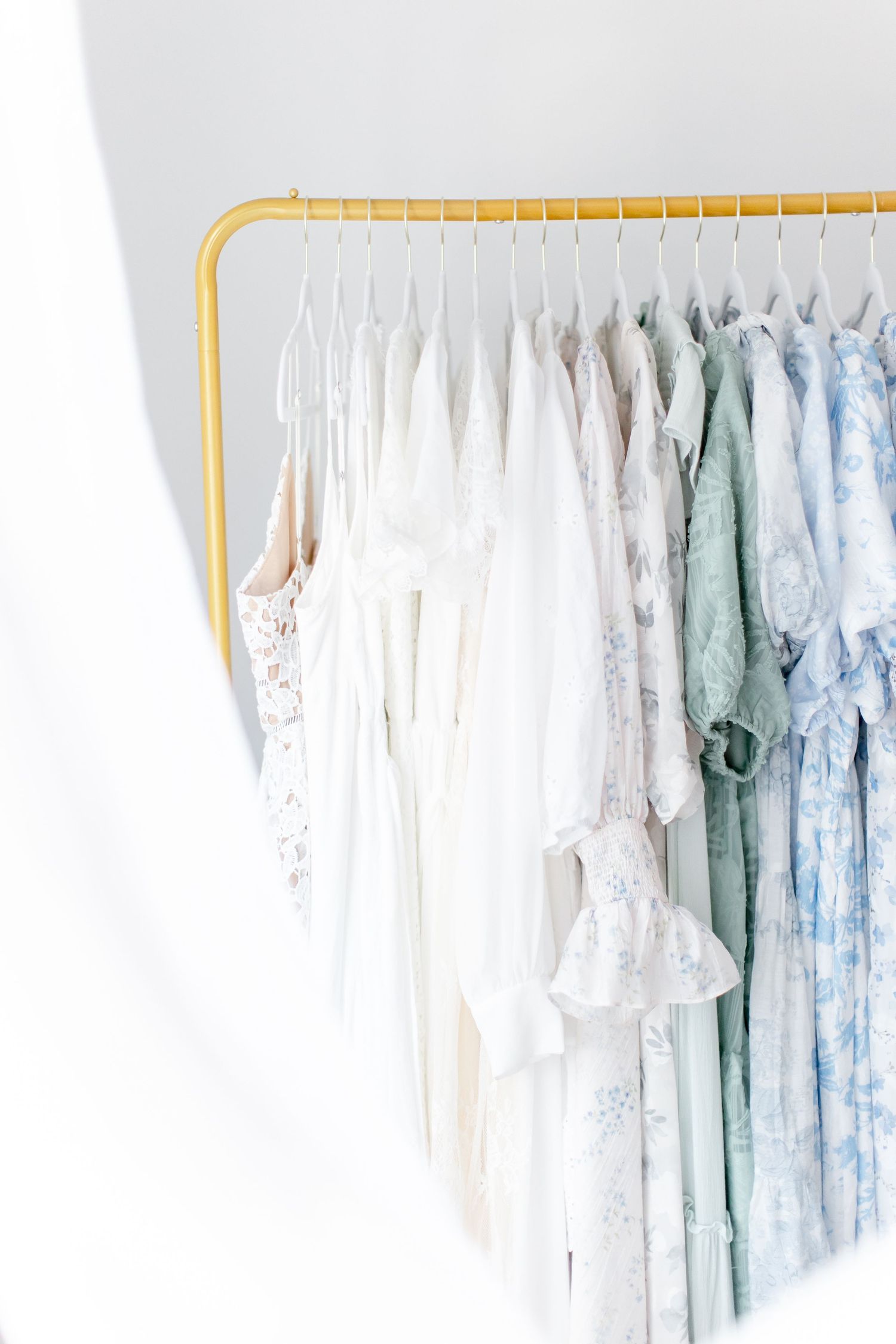 Delicate white and pastel dresses hanging on a gold clothing rack against a white wall in an ombre color arrangement.