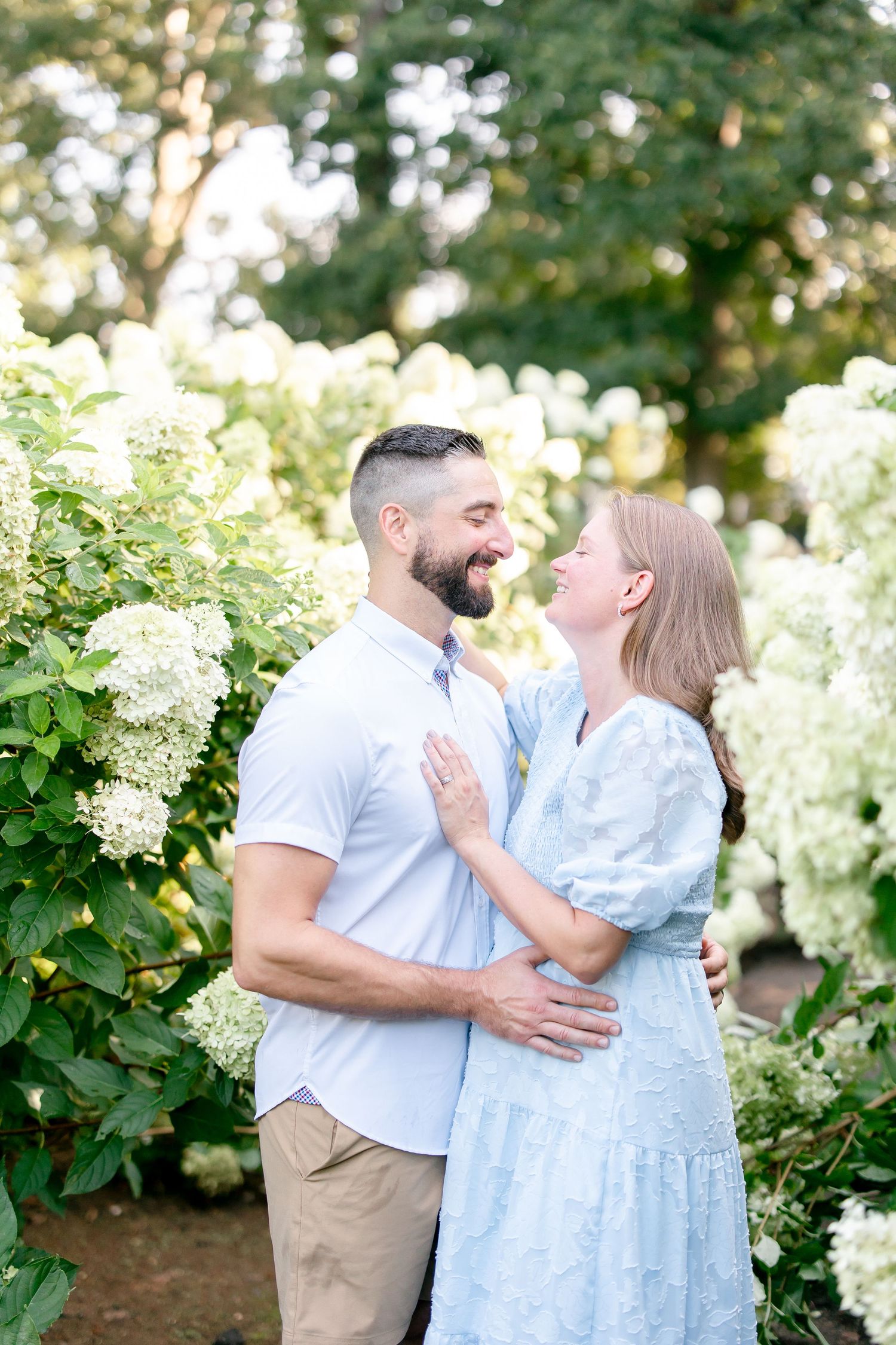 Couple shares loving embrace among blooming white hydrangea bushes in garden setting.
