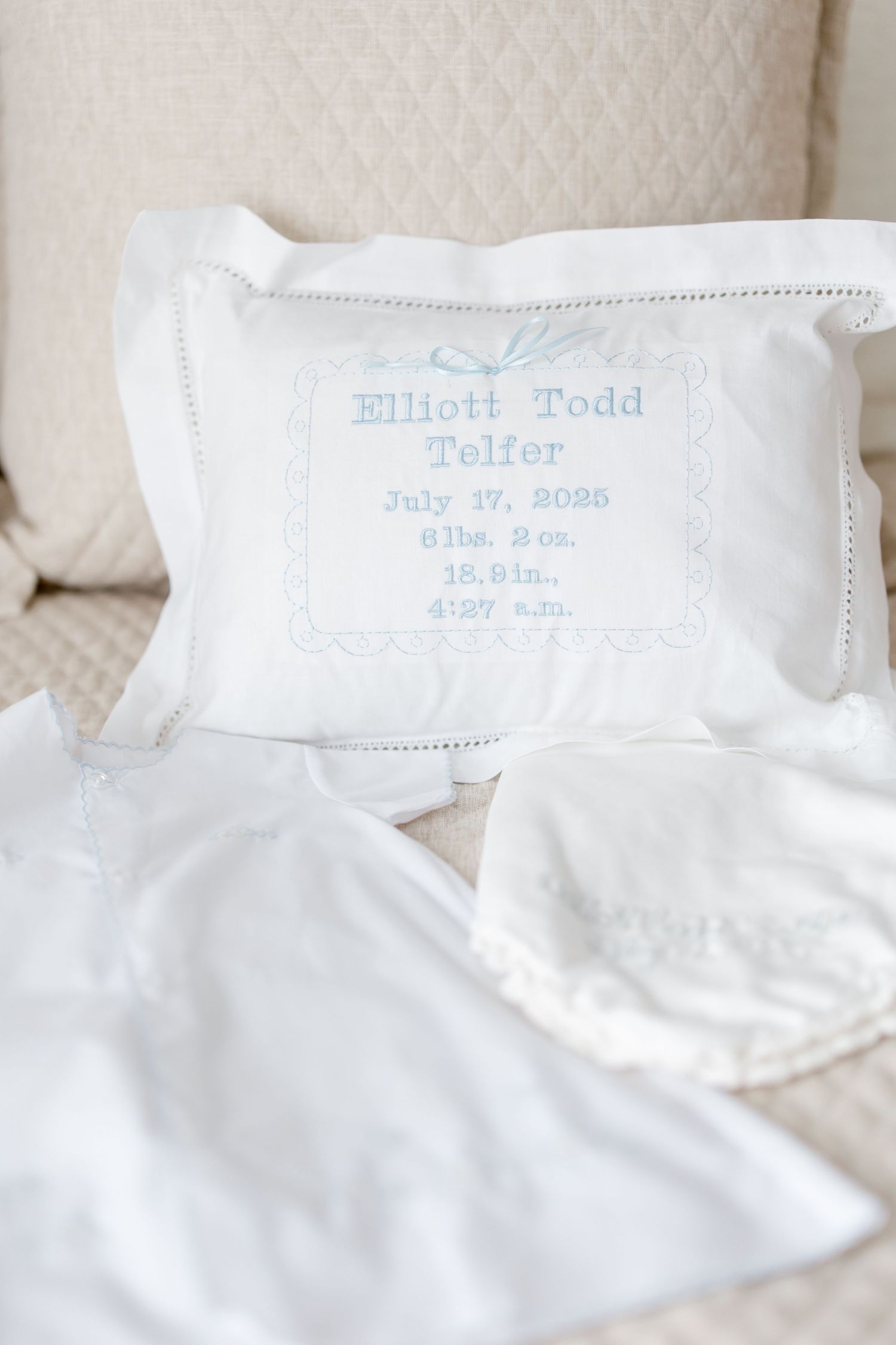 Embroidered white baby garments with blue cursive text laying on light colored bedding.