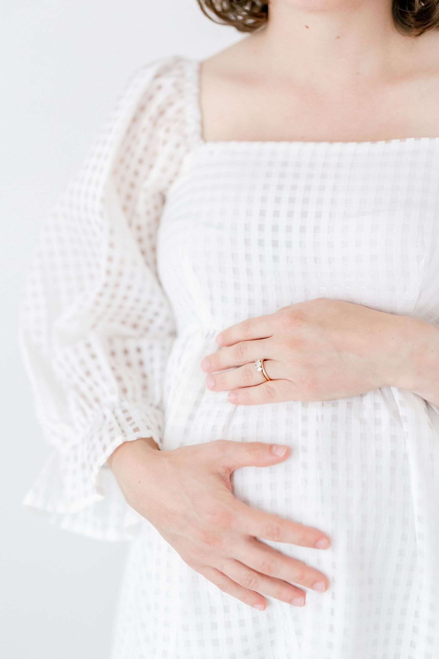 Woman in white knit sweater shows delicate engagement ring on her hand against light background.