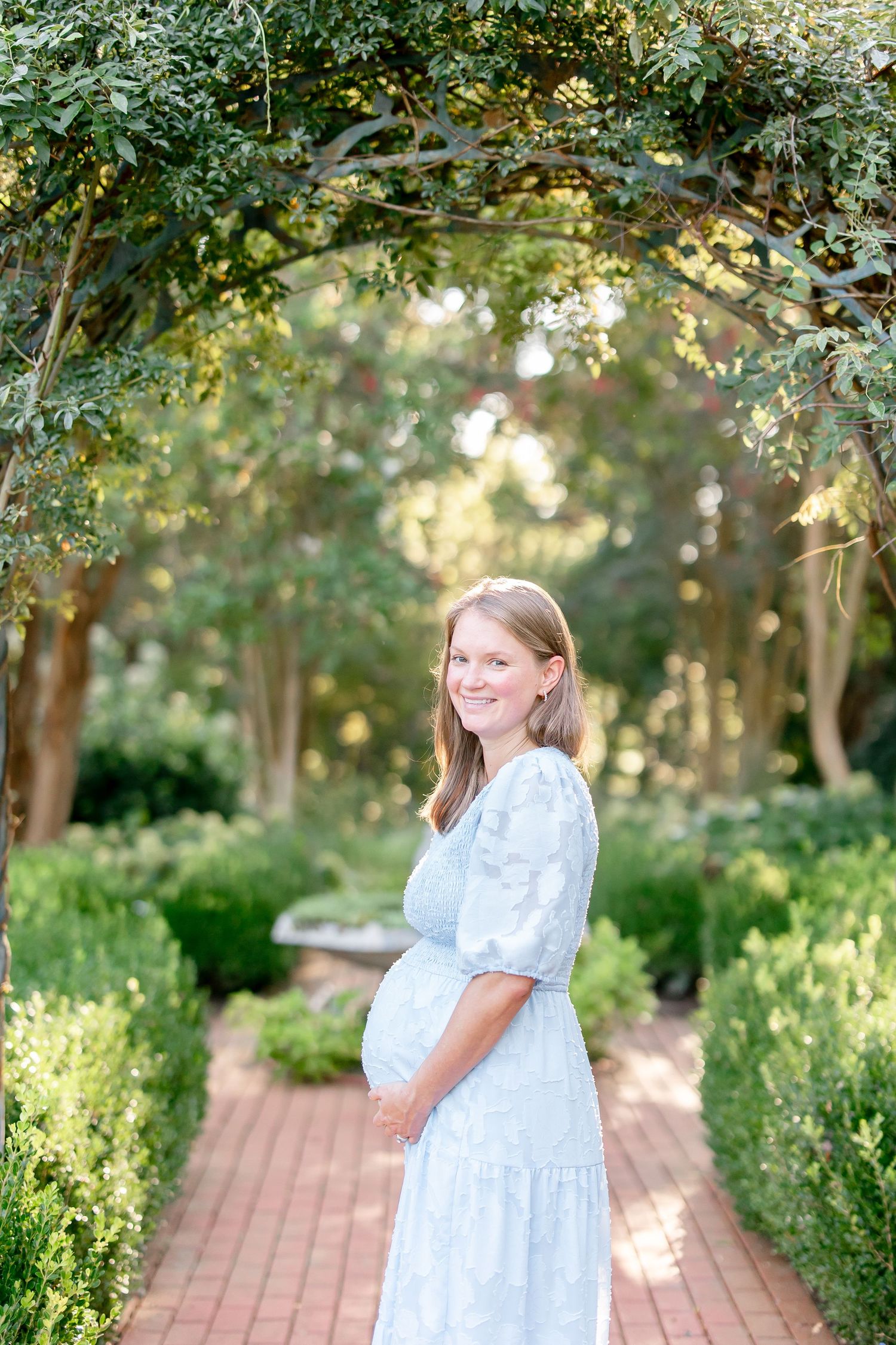 Woman in light blue dress stands on brick path surrounded by lush garden landscaping.