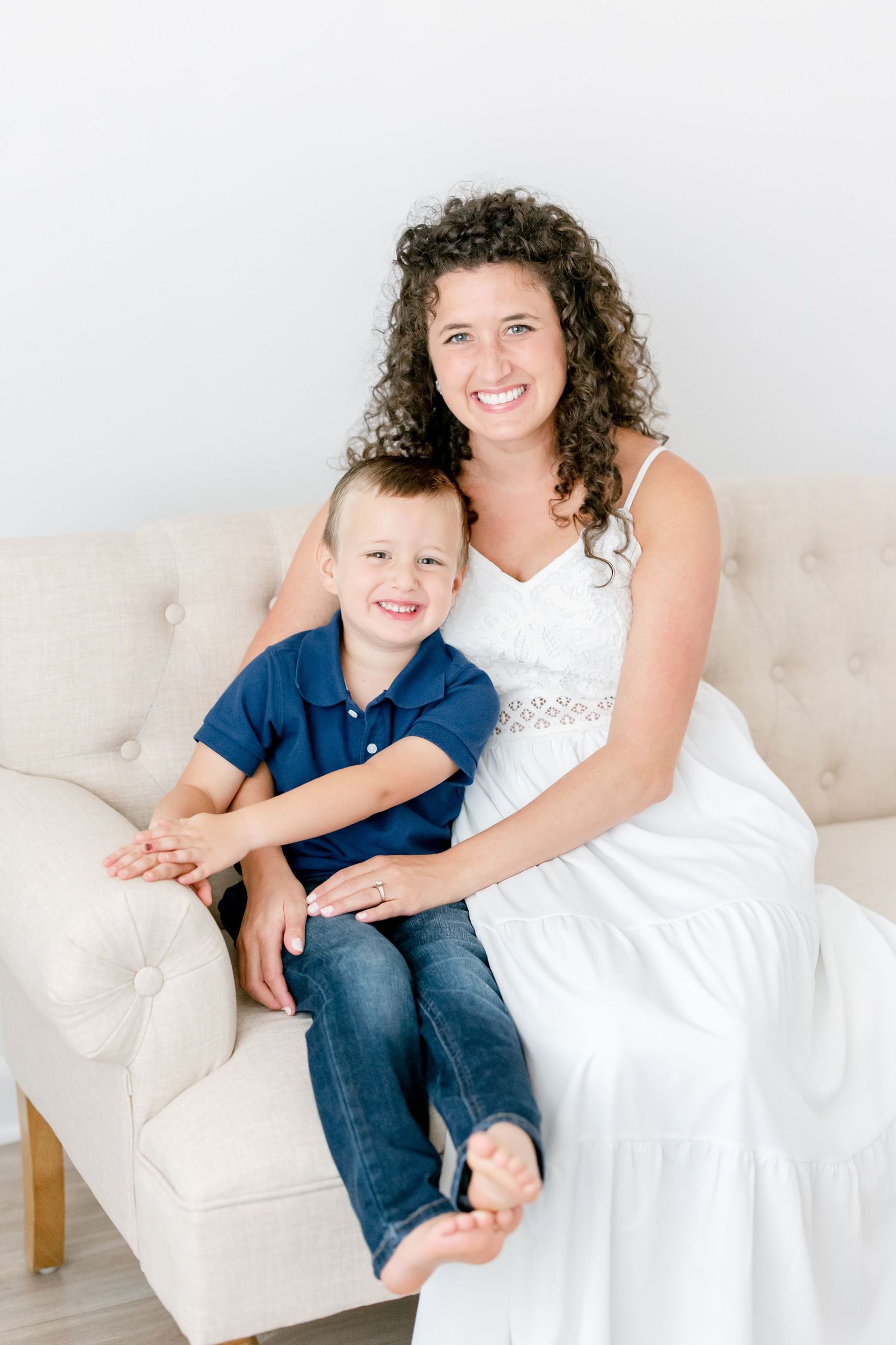 Mother in white dress and child in blue share loving moment on light couch against white backdrop.
