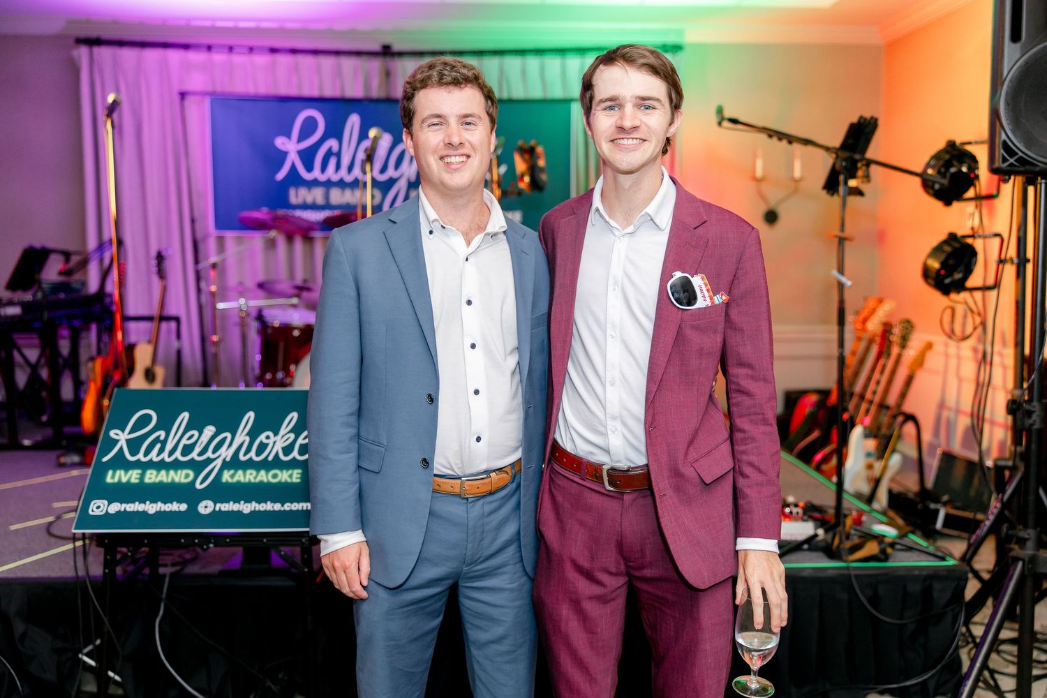Two performers pose in suits in front of Raleighoke branded backdrop with stage equipment visible.