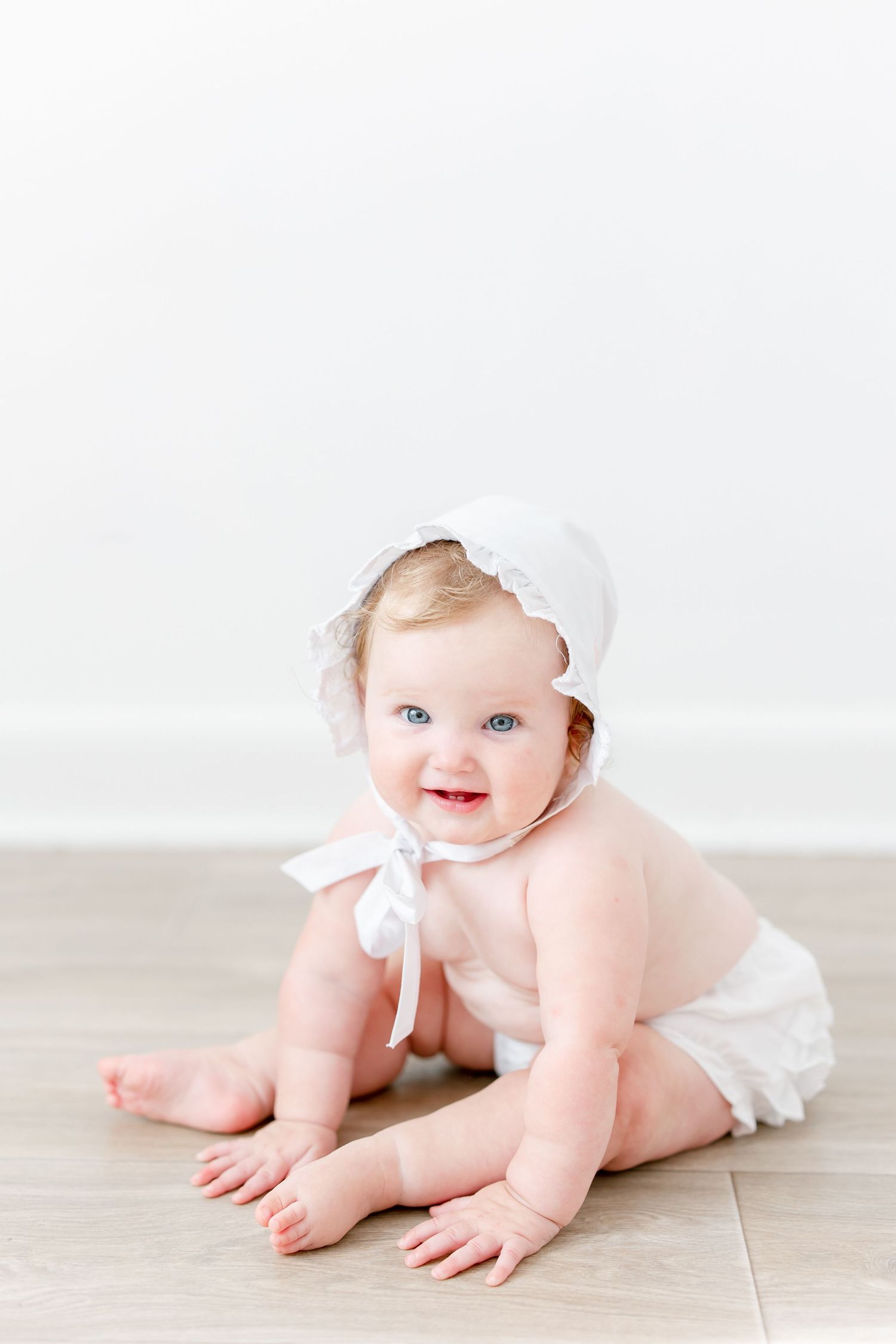 Baby in white bonnet and diaper crawls on light wooden floor in minimalist studio setting.