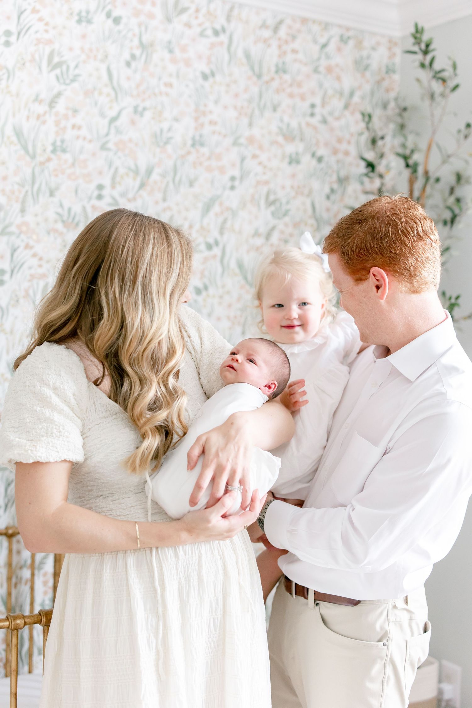 Parents in white outfits share tender moments with their baby against floral wallpaper in series of candid photos.
