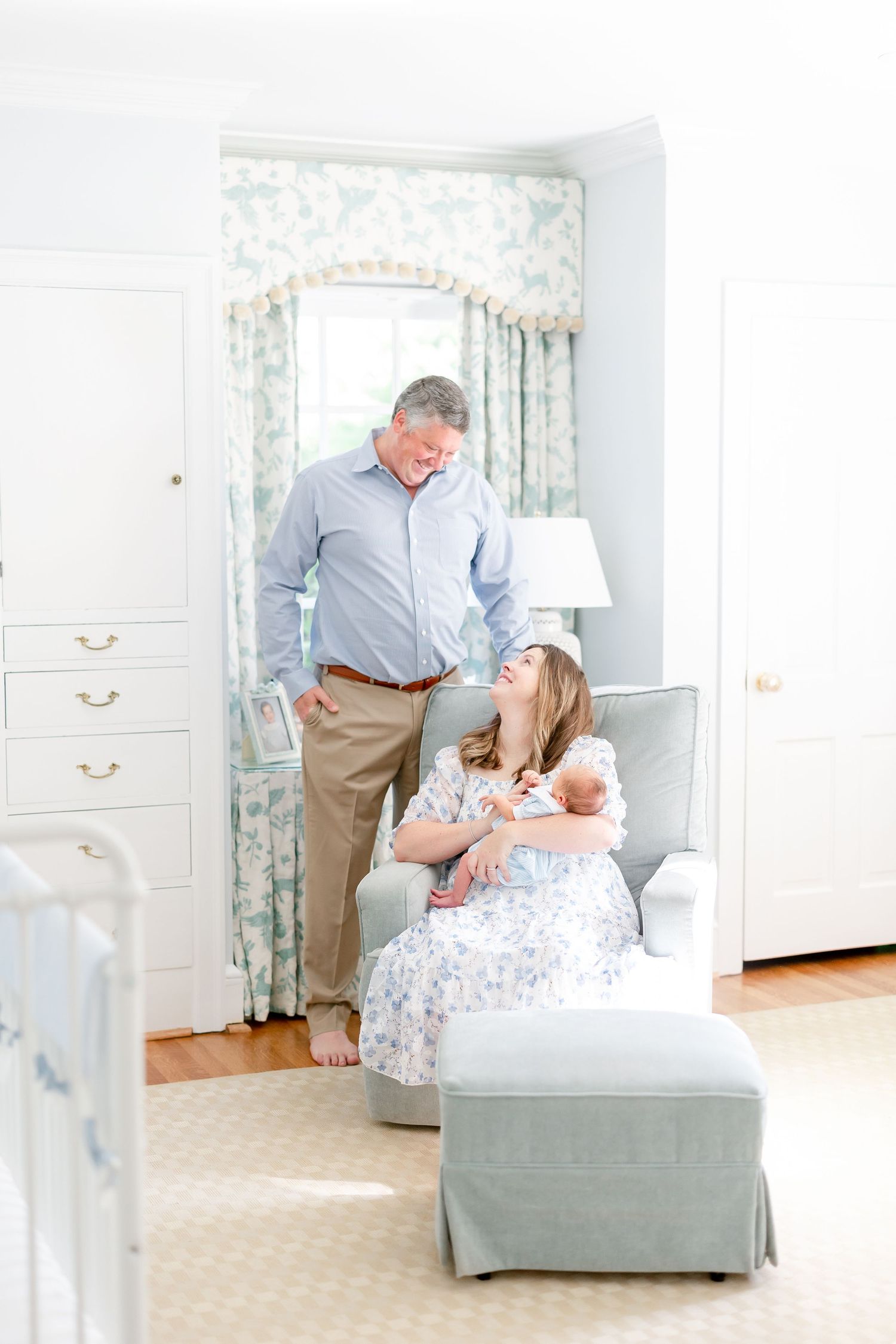 New parents admire their newborn baby in a serene nursery, mother seated in a gray glider chair holding the infant.