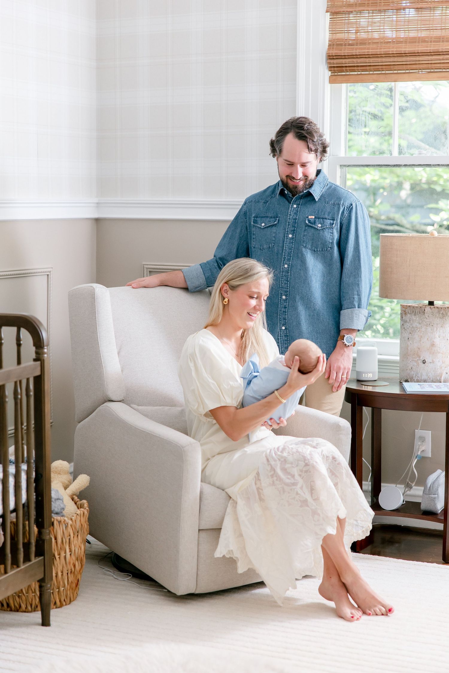 A family sits together on a white armchair in a bright nursery room with bamboo shades and wooden accents.