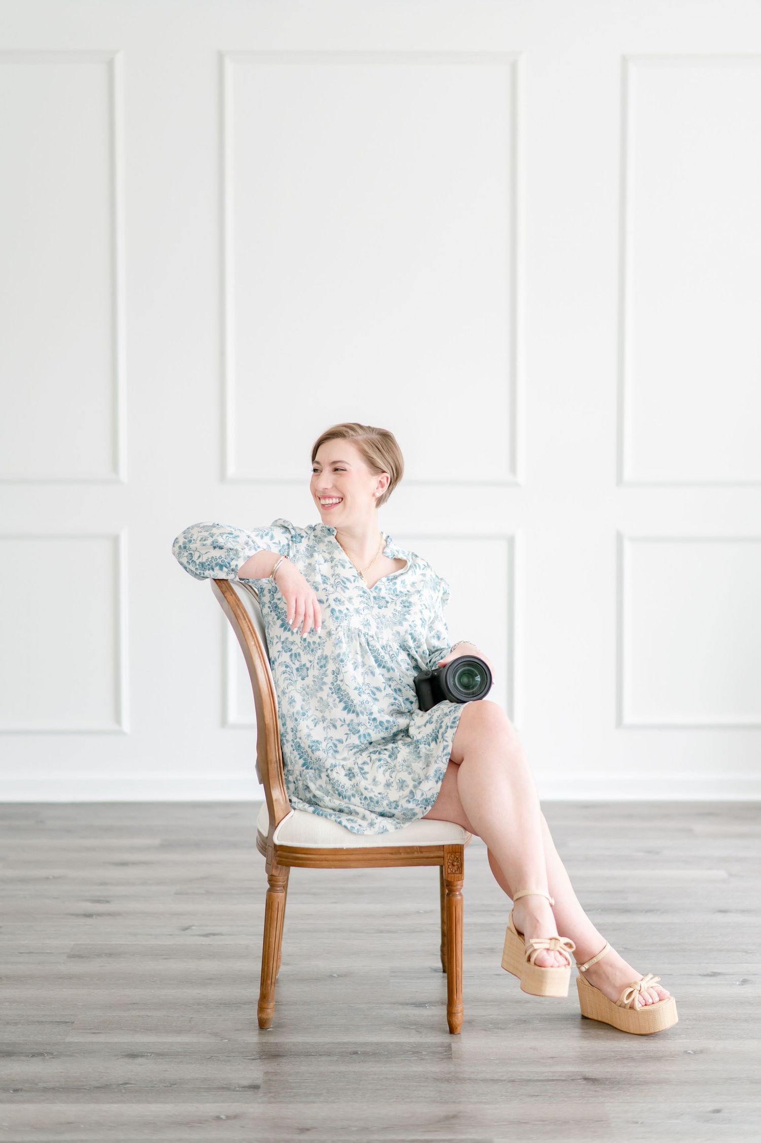 Professional photographer in floral dress sits casually on wooden chair in bright white studio with camera in hand.