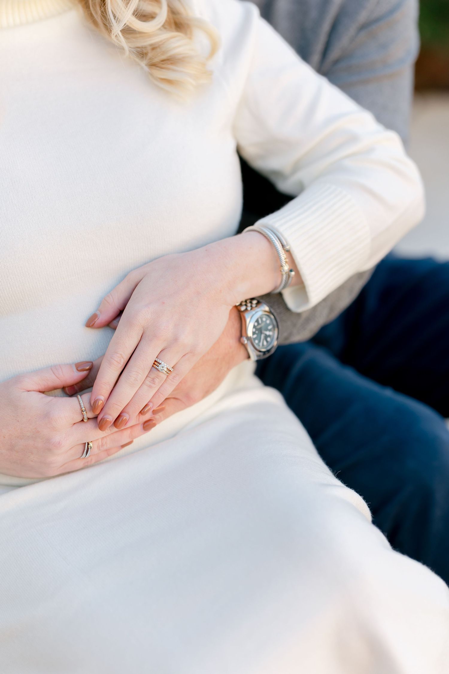 Woman in a cream knit sweater and skirt cradling her baby bump, wearing rings and a silver watch, with a partner's hand resting on top.