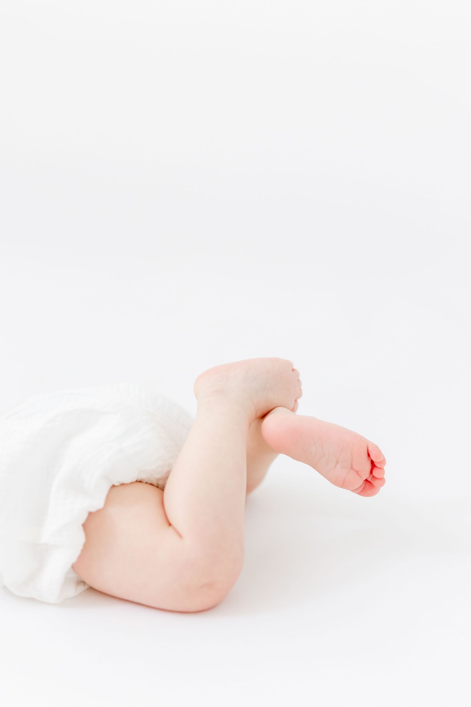 Close-up of a baby's chubby legs and tiny bare feet in a white diaper, resting against a bright, clean white background.