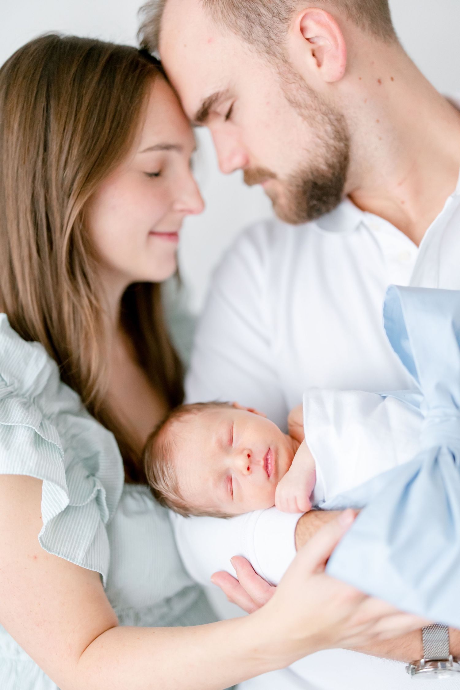 A sleeping newborn baby swaddled in white and blue is tenderly held by parents in an intimate family portrait.