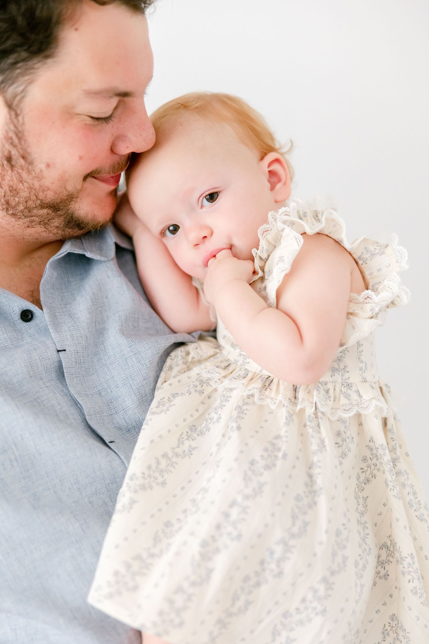 A tender parent-child moment captured in soft light as the baby wears a delicate cream-colored lace dress.