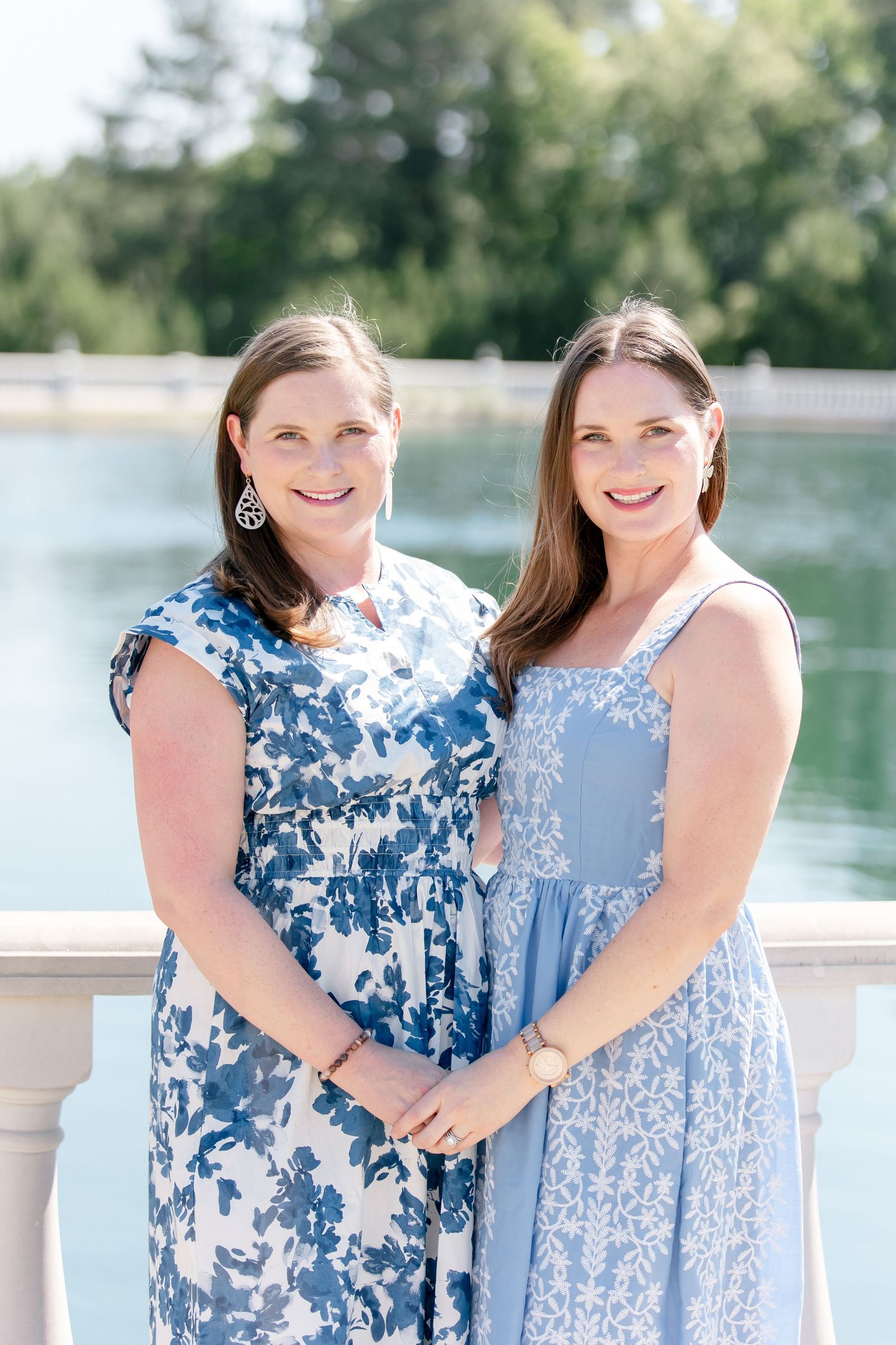 Two women in coordinating blue and white floral dresses pose together by a water feature on a sunny day.