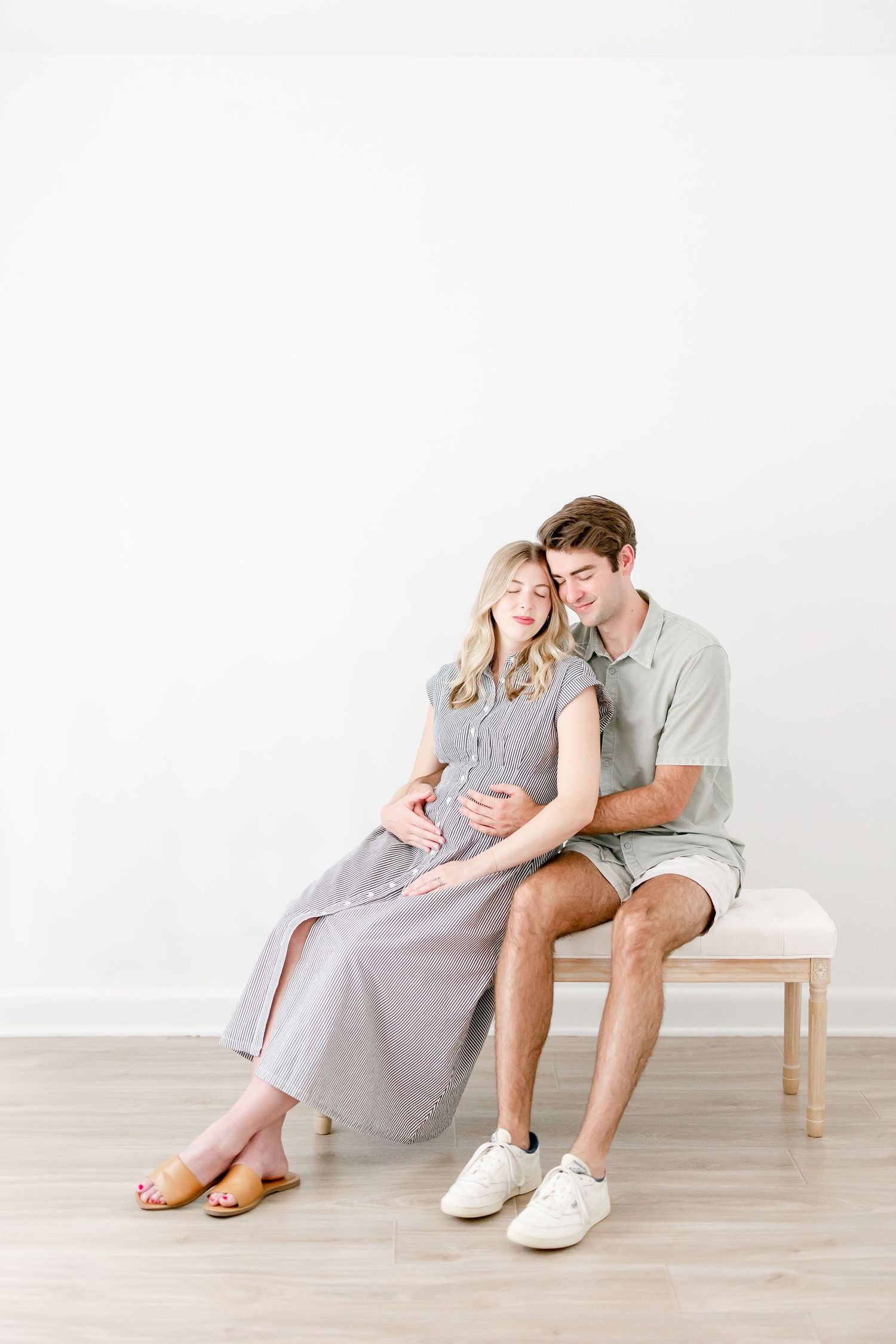 Couple relaxes on wooden bench in casual attire during maternity photo session.