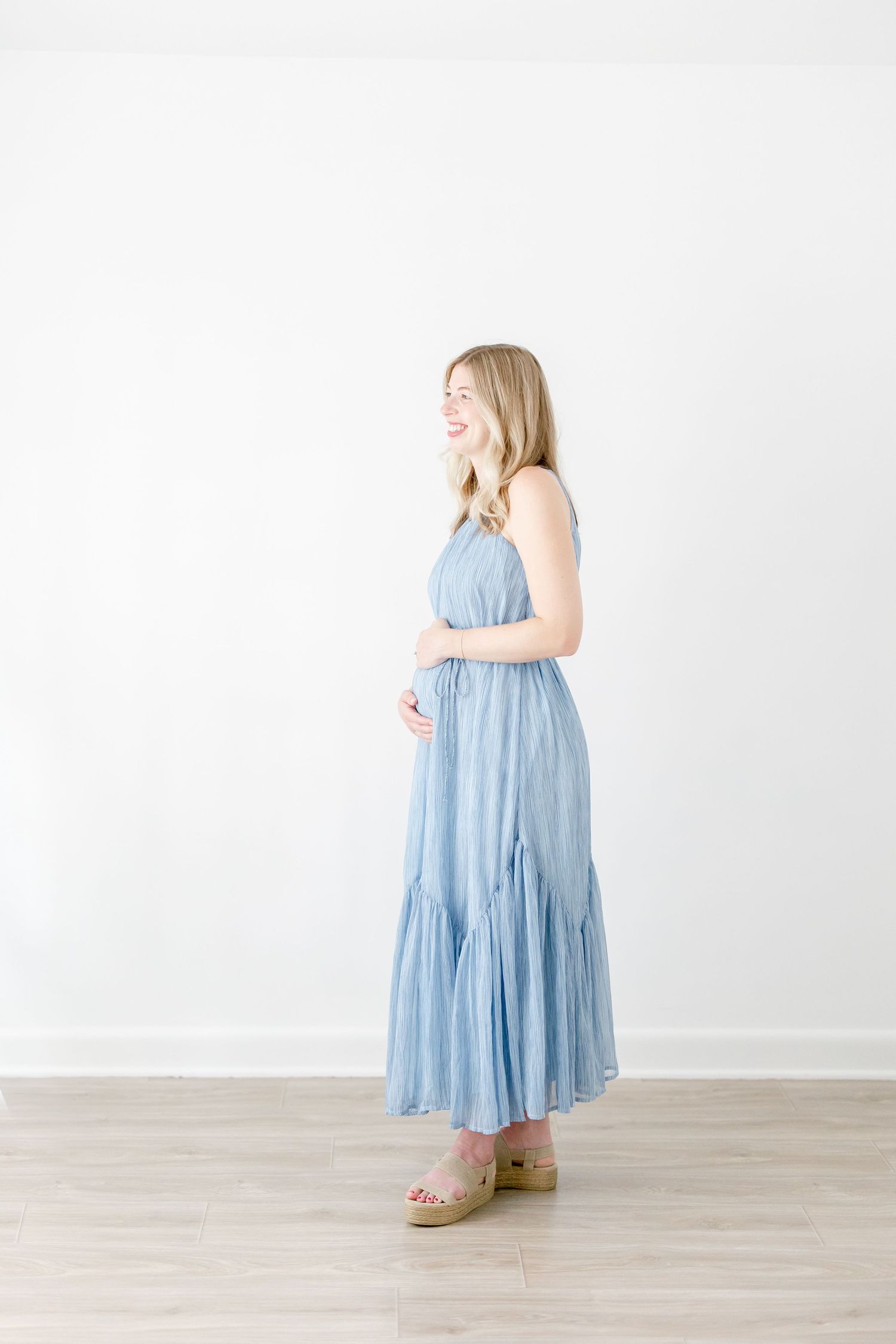 Pregnant woman in flowing light blue midi dress poses against white backdrop in four different angles.