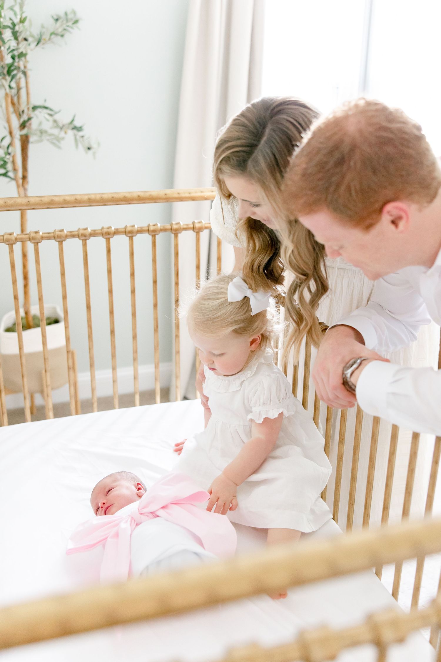 Family gathers around wooden crib in nursery with natural light streaming through sheer curtains.