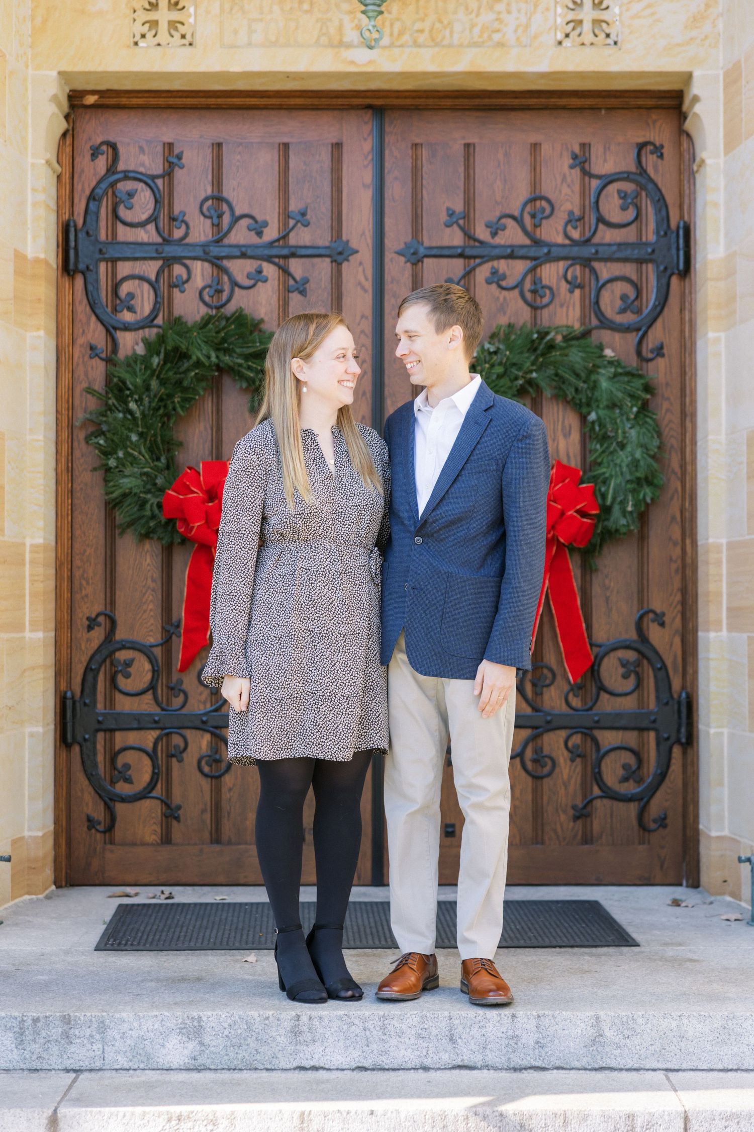 Couple in winter attire standing in front of ornate iron church doors decorated with holiday wreaths and red bows.