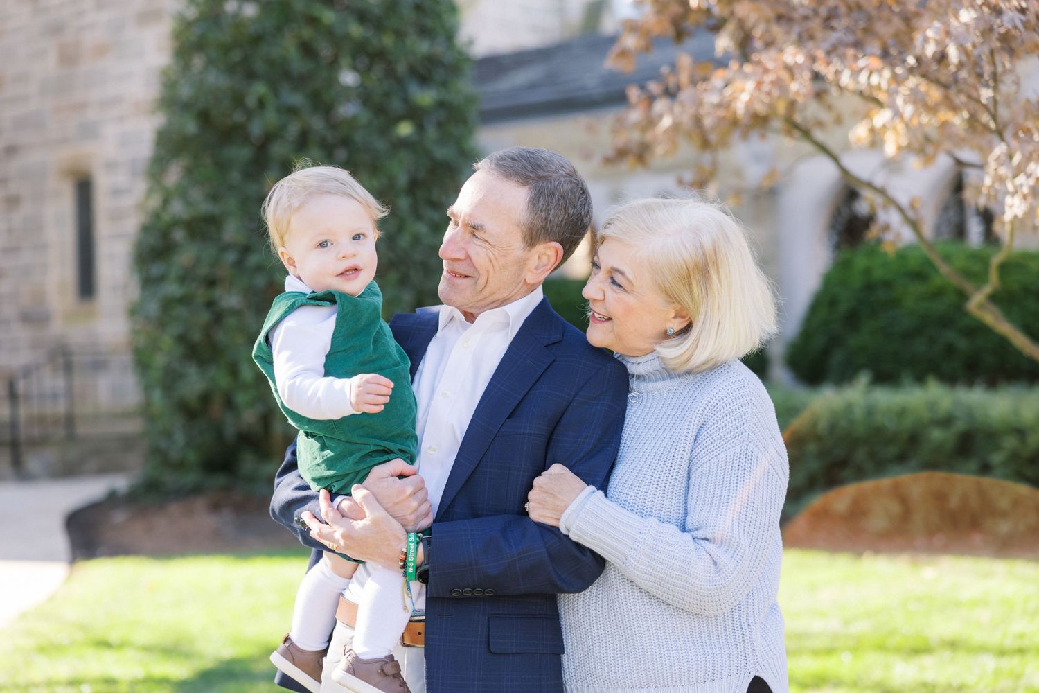 A grandparent couple wearing business casual attire stands outdoors on a sunny day with a small child.