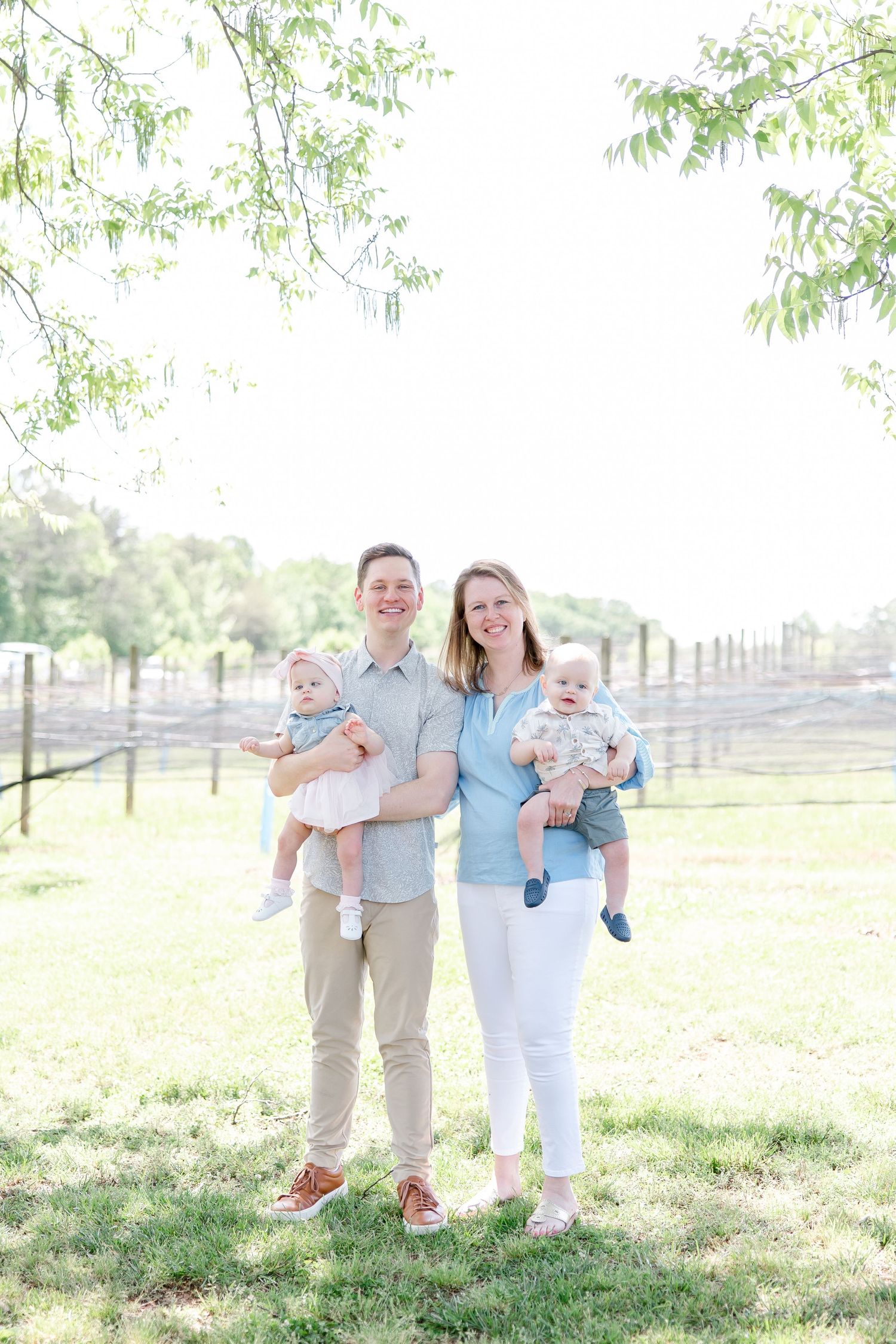 Family portrait in a pastoral setting with sunlit trees and fencing in the background.