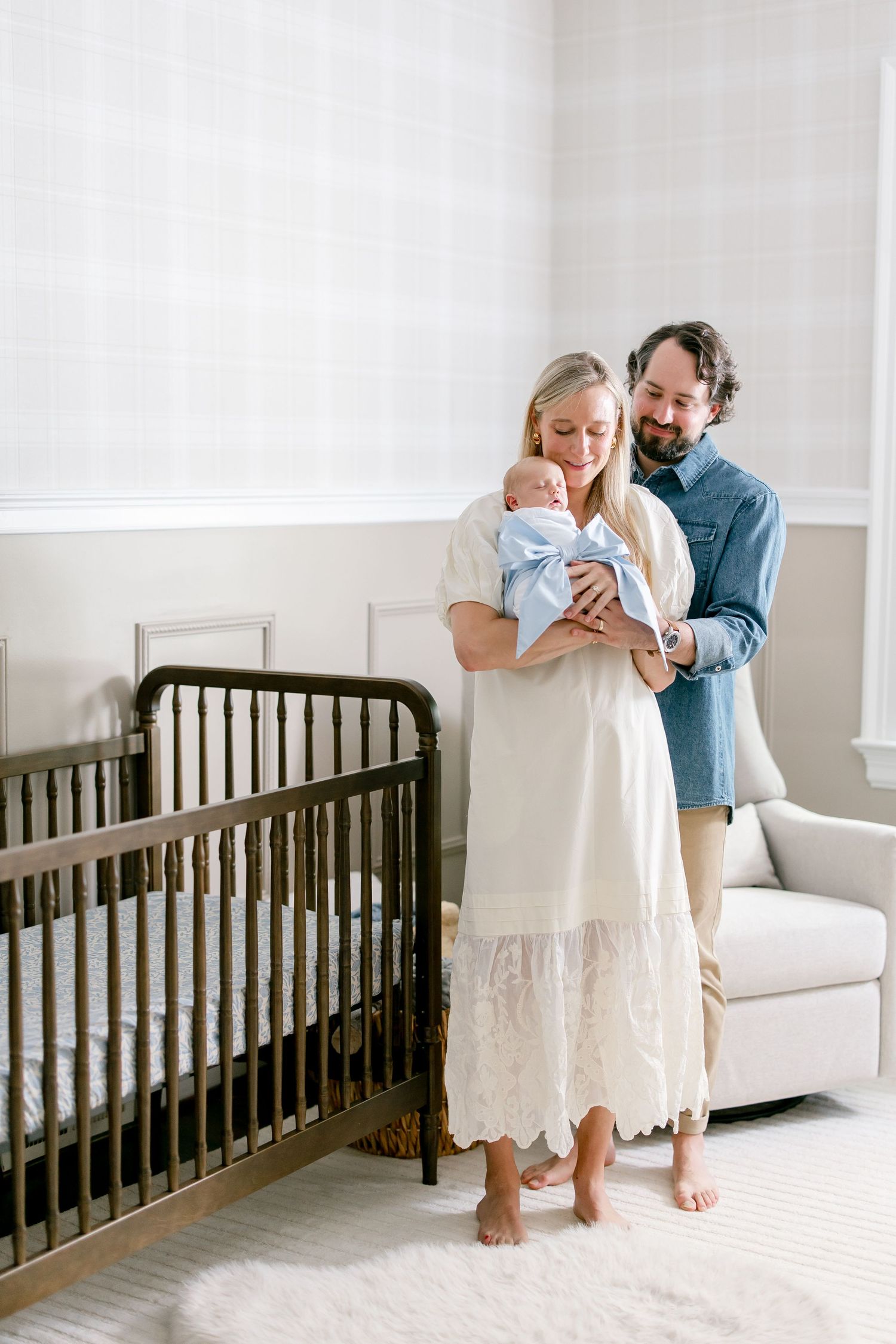 Parents sharing a tender moment with their baby in a minimalist white nursery with dark wood crib.