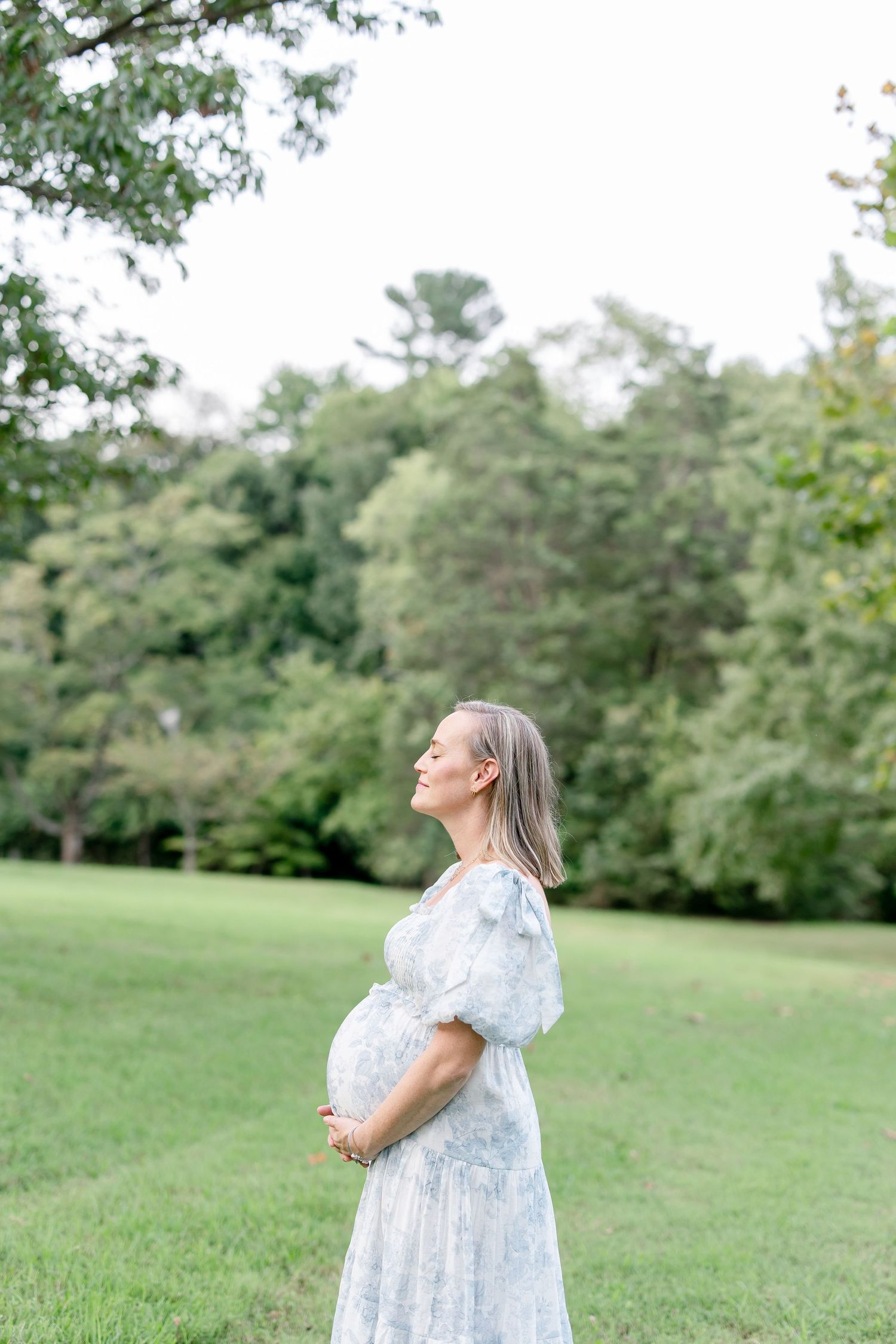 A pregnant woman in a light blue floral dress stands sideways in a peaceful garden setting.