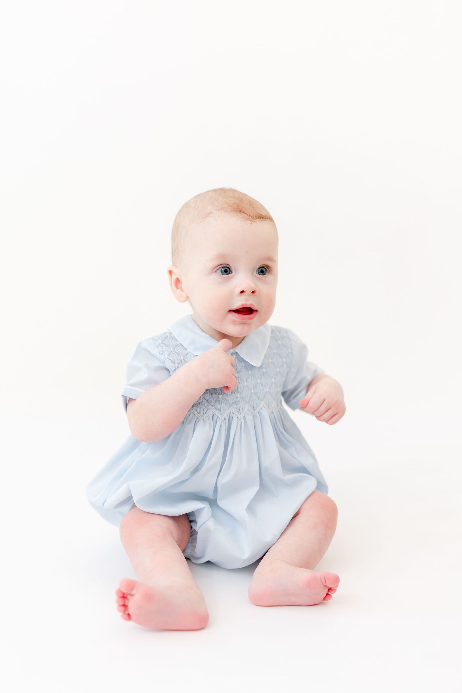 Baby sitting barefoot on a white background wearing a light blue smocked romper with a white collar and short sleeves.