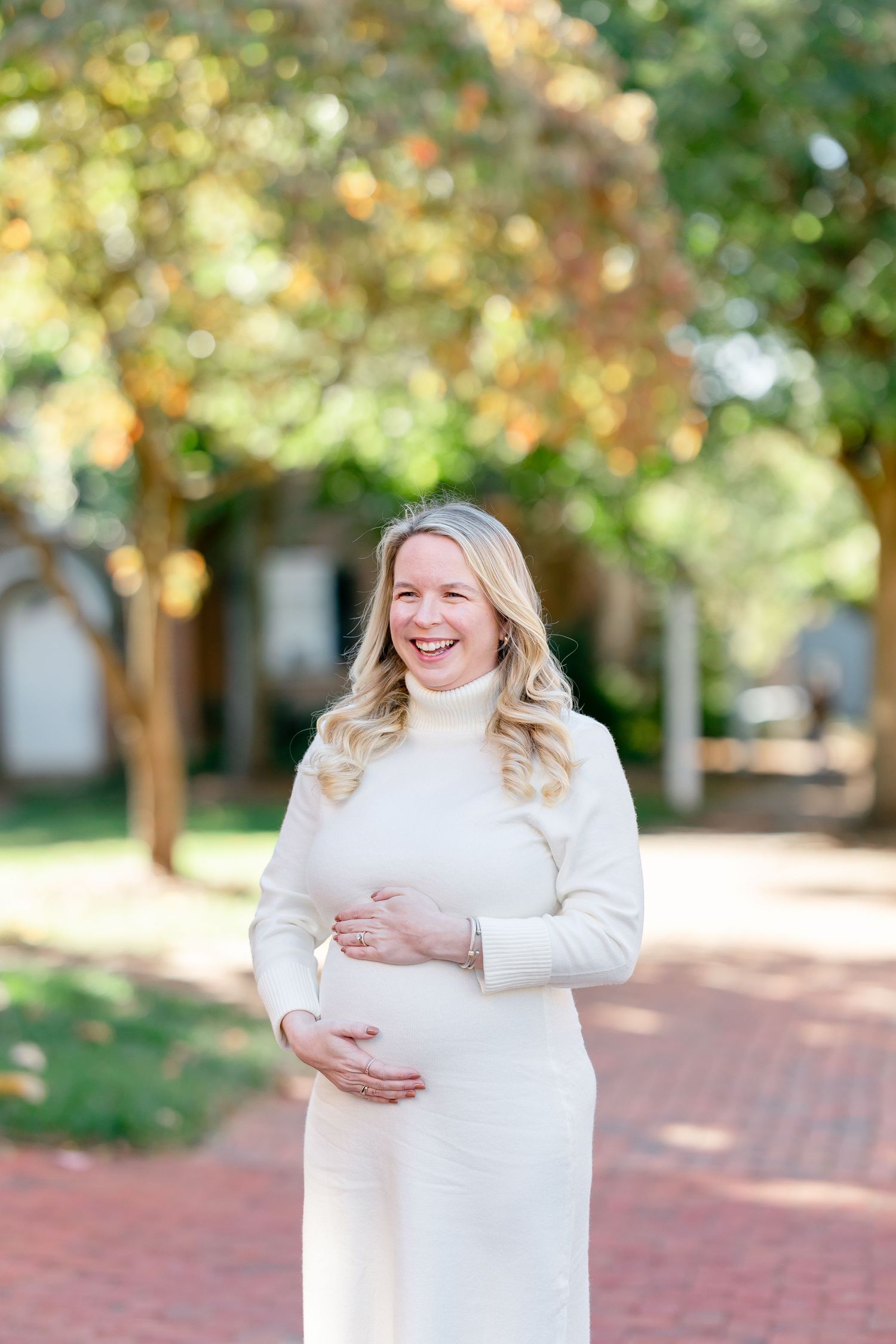 Smiling pregnant woman in a cream knit turtleneck dress cradles her belly on a sunlit brick path lined with autumn trees.