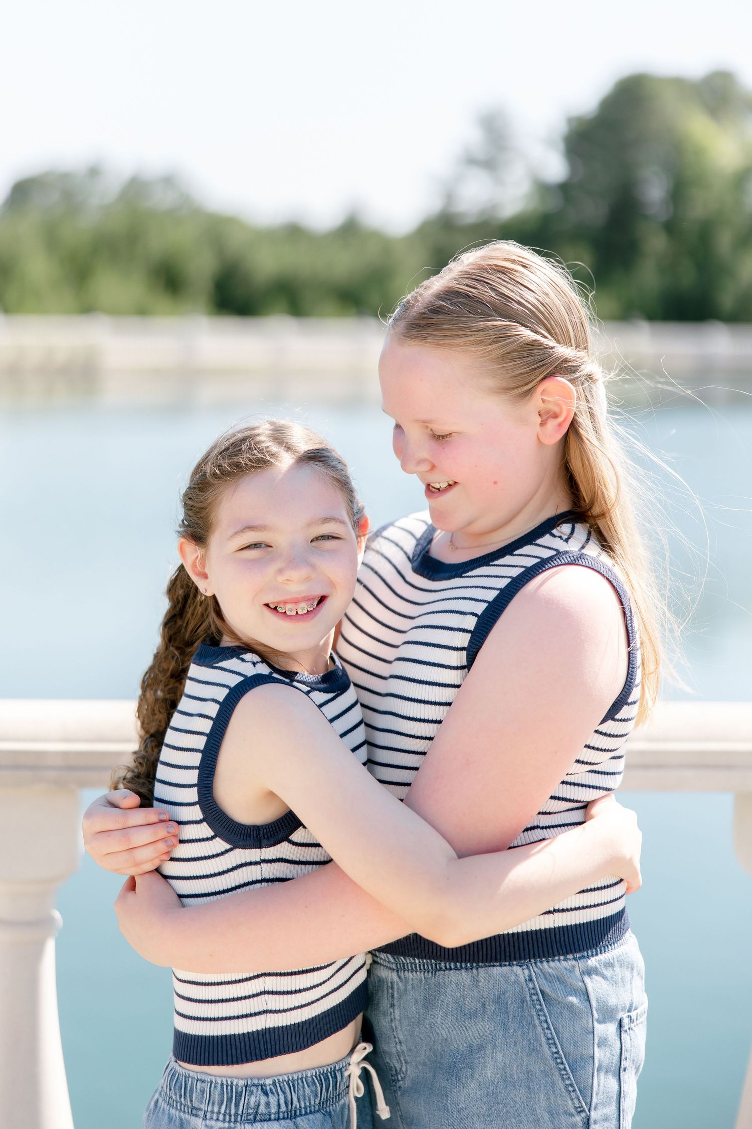 Two people in matching striped sleeveless tops embrace and smile by a waterfront on a sunny day.