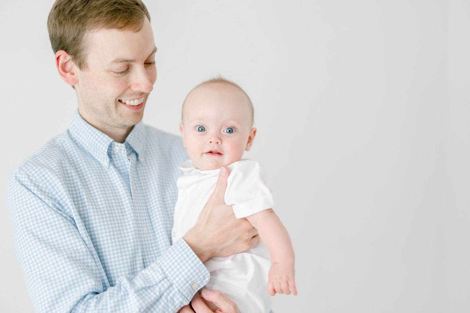 A parent in a blue checkered shirt holds a baby wearing white against a light background.