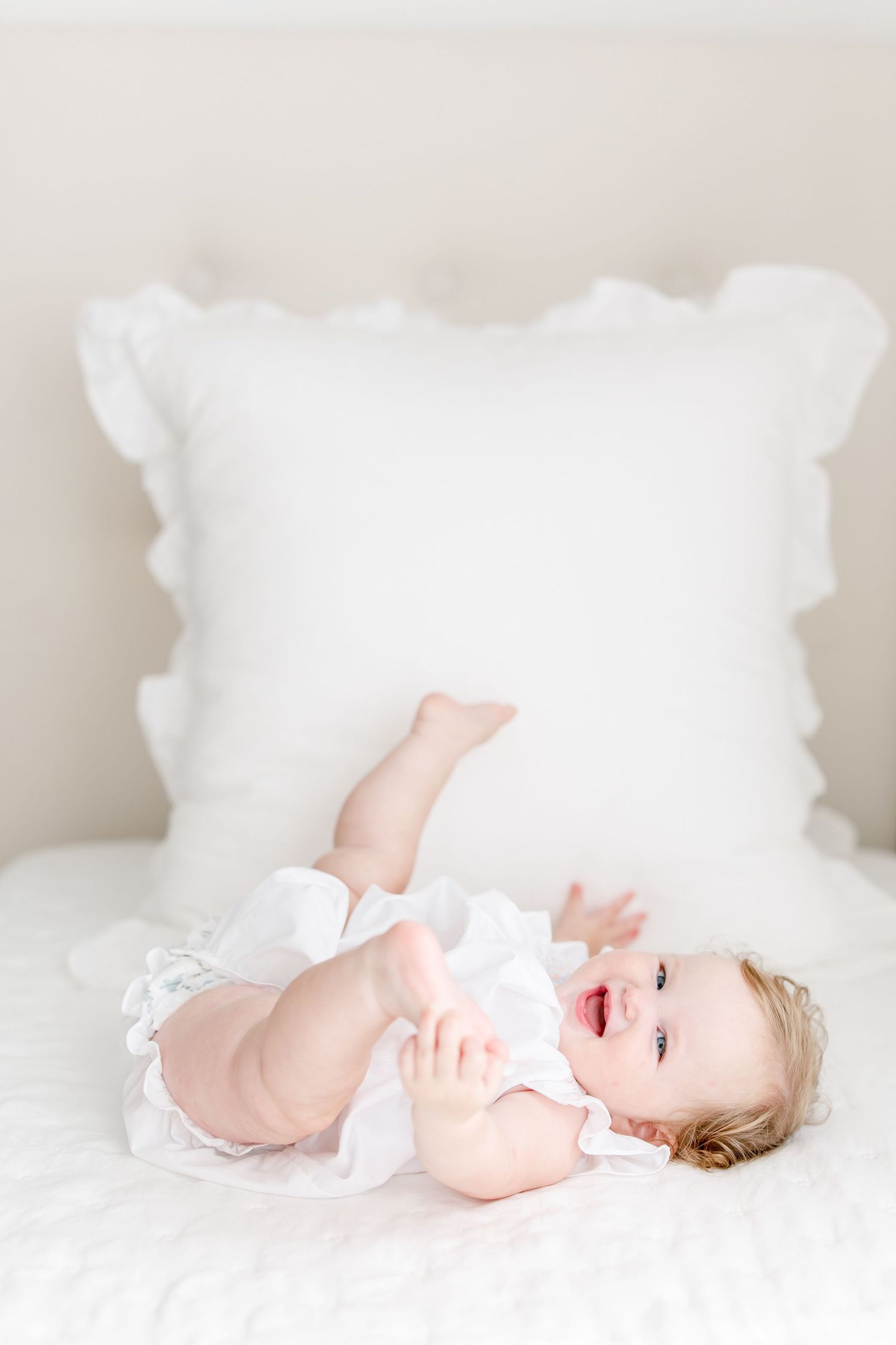 Laughing baby wearing white dress lying on cozy white bedding with decorative ruffled pillow in background.
