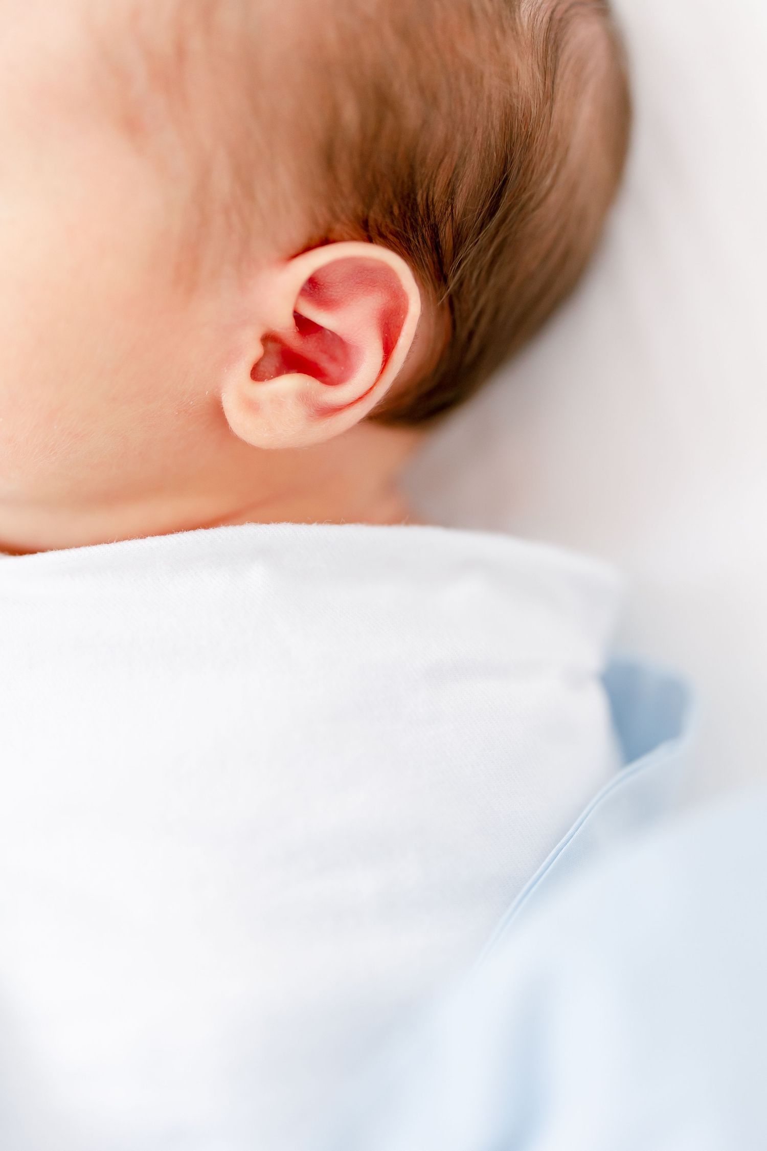Close-up of a sleeping baby's ear and head against white bedding, wearing light blue clothing.
