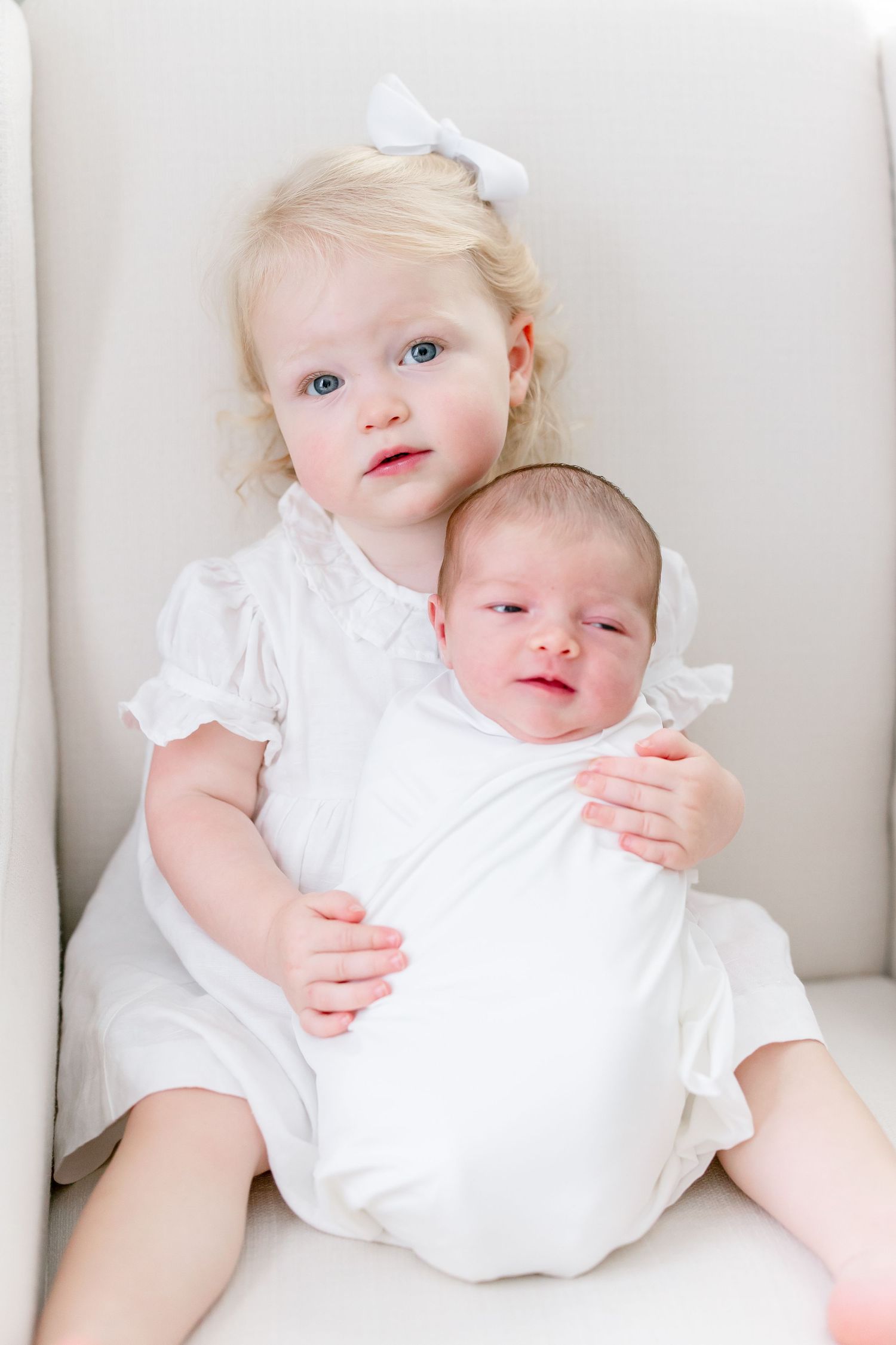 An infant wearing a white dress rests on plush white furniture in a bright room.