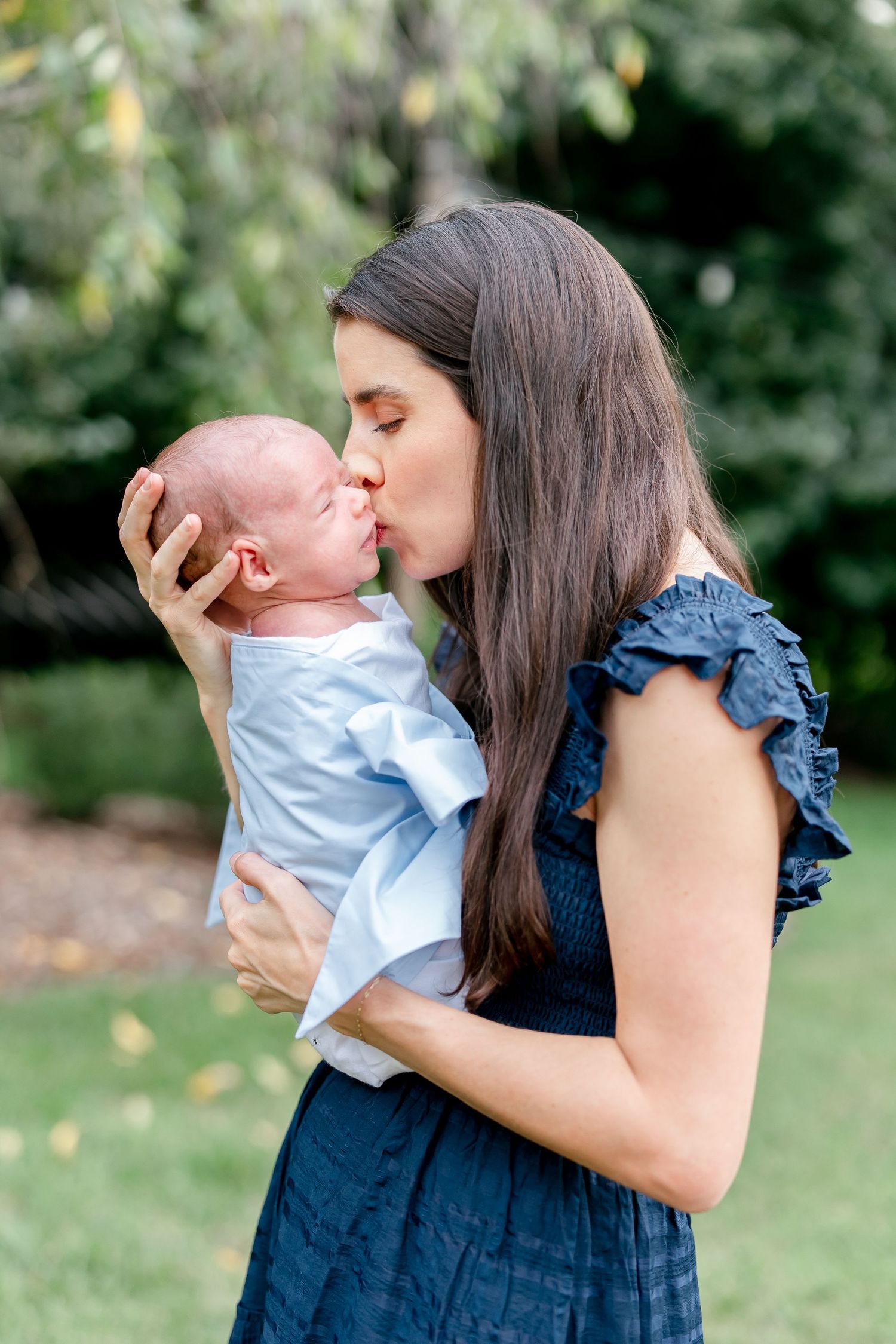 Mother in navy dress holding and kissing newborn baby in light blue outfit outdoors.