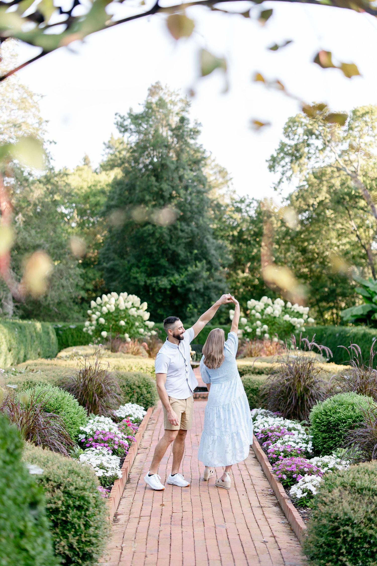 Two people dancing and twirling together on a brick pathway amidst flowering garden plants.