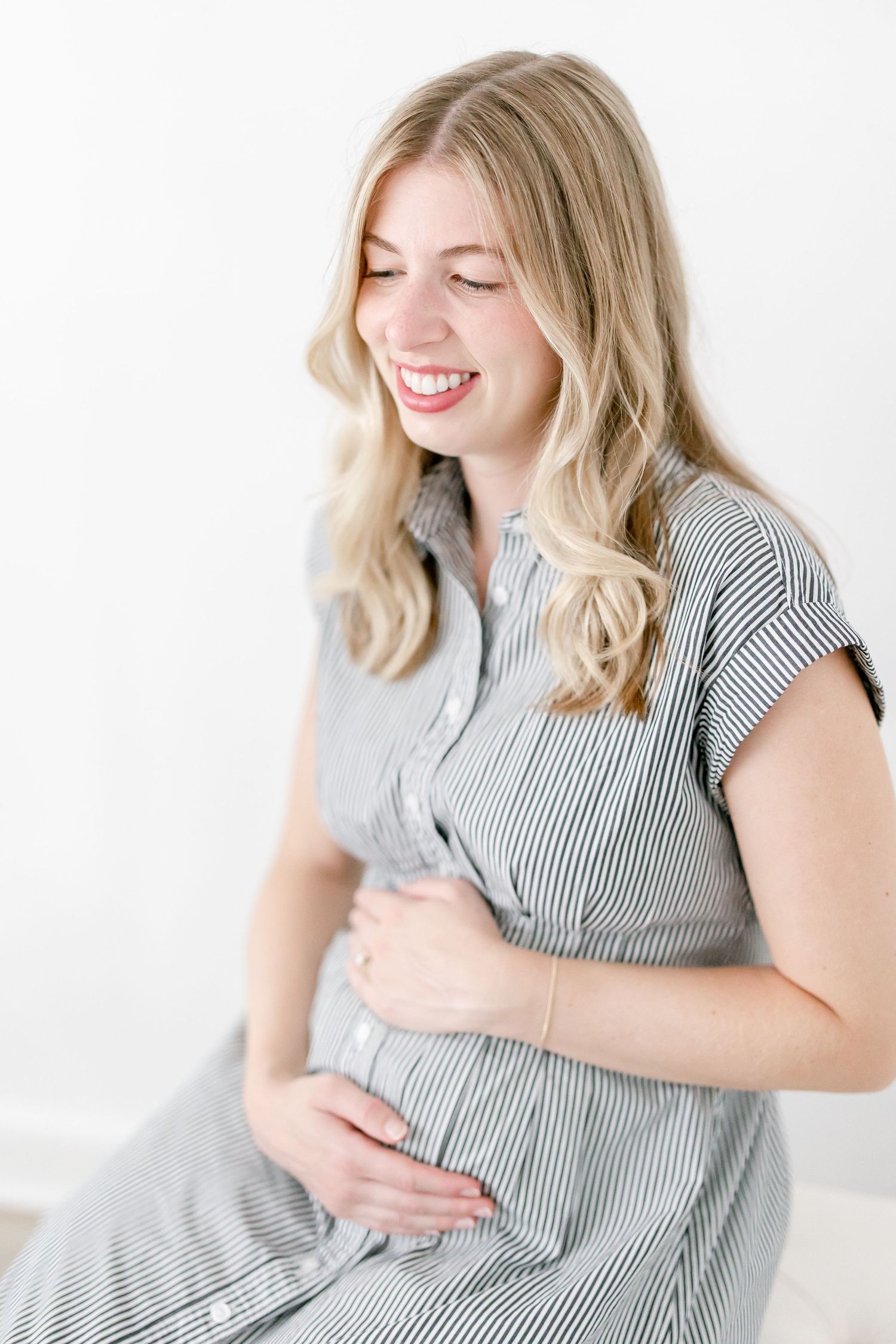 Woman in striped dress smiling joyfully while holding her pregnant belly against white background.