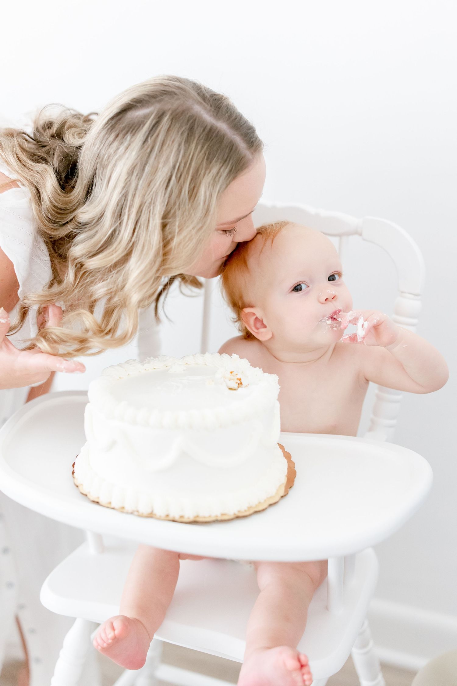 Baby having fun while eating cake and smashing it with their hands on a white highchair in a bright room.