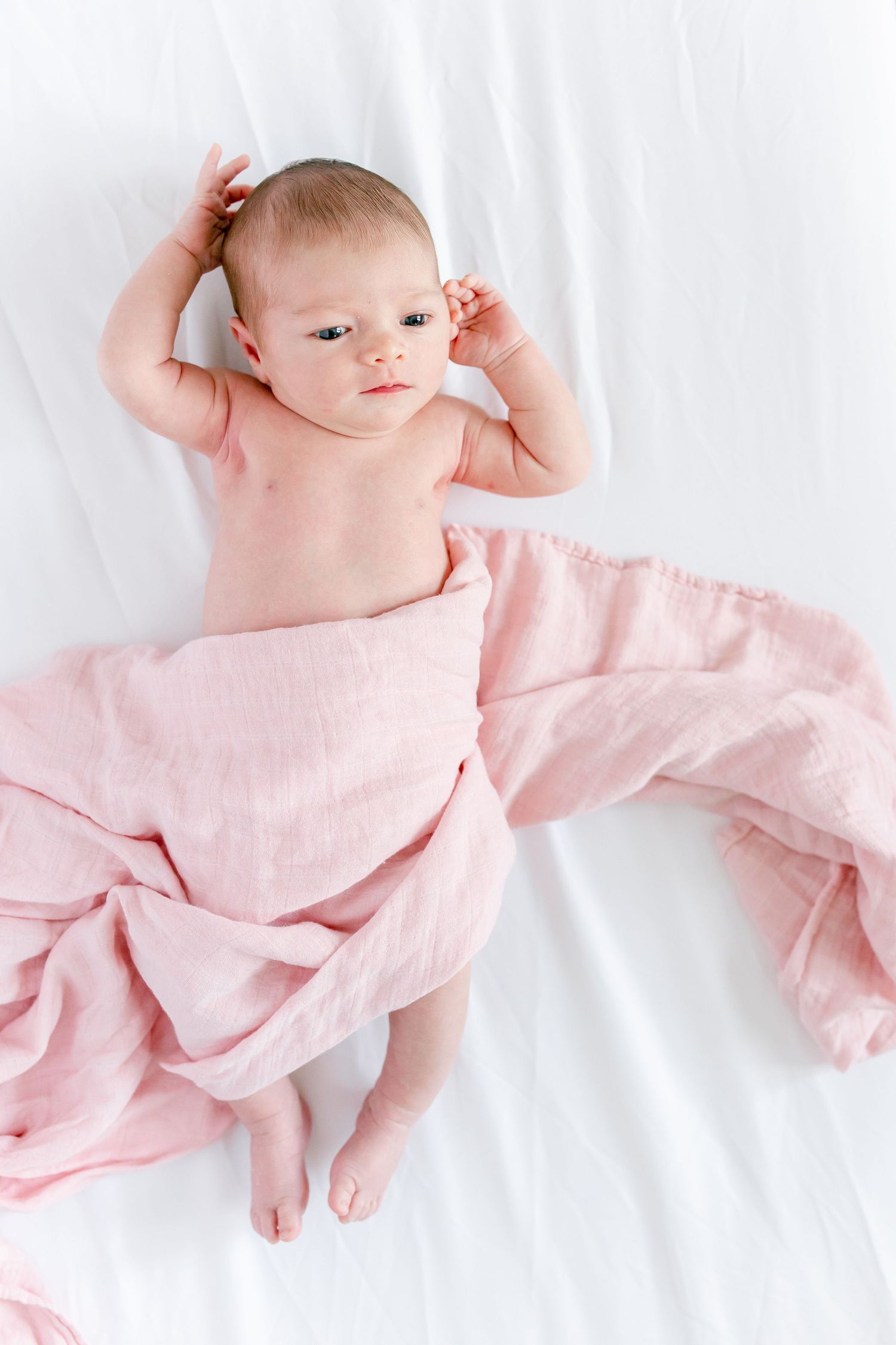 A baby lying on white bedding wrapped in soft pink fabric while stretching.