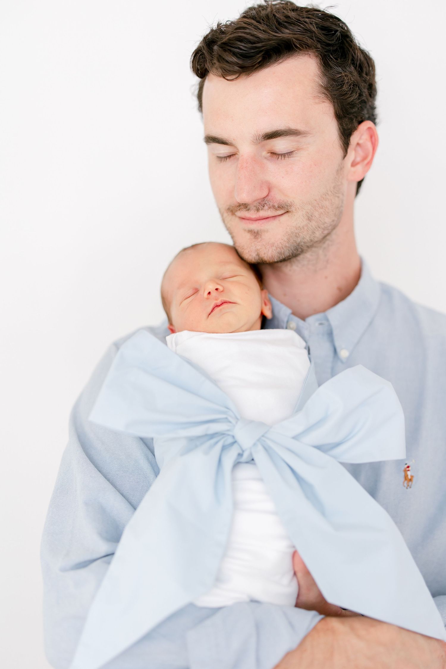 A tender moment shows a sleeping newborn wrapped in a light blue bow blanket being held against a blue button-down shirt.