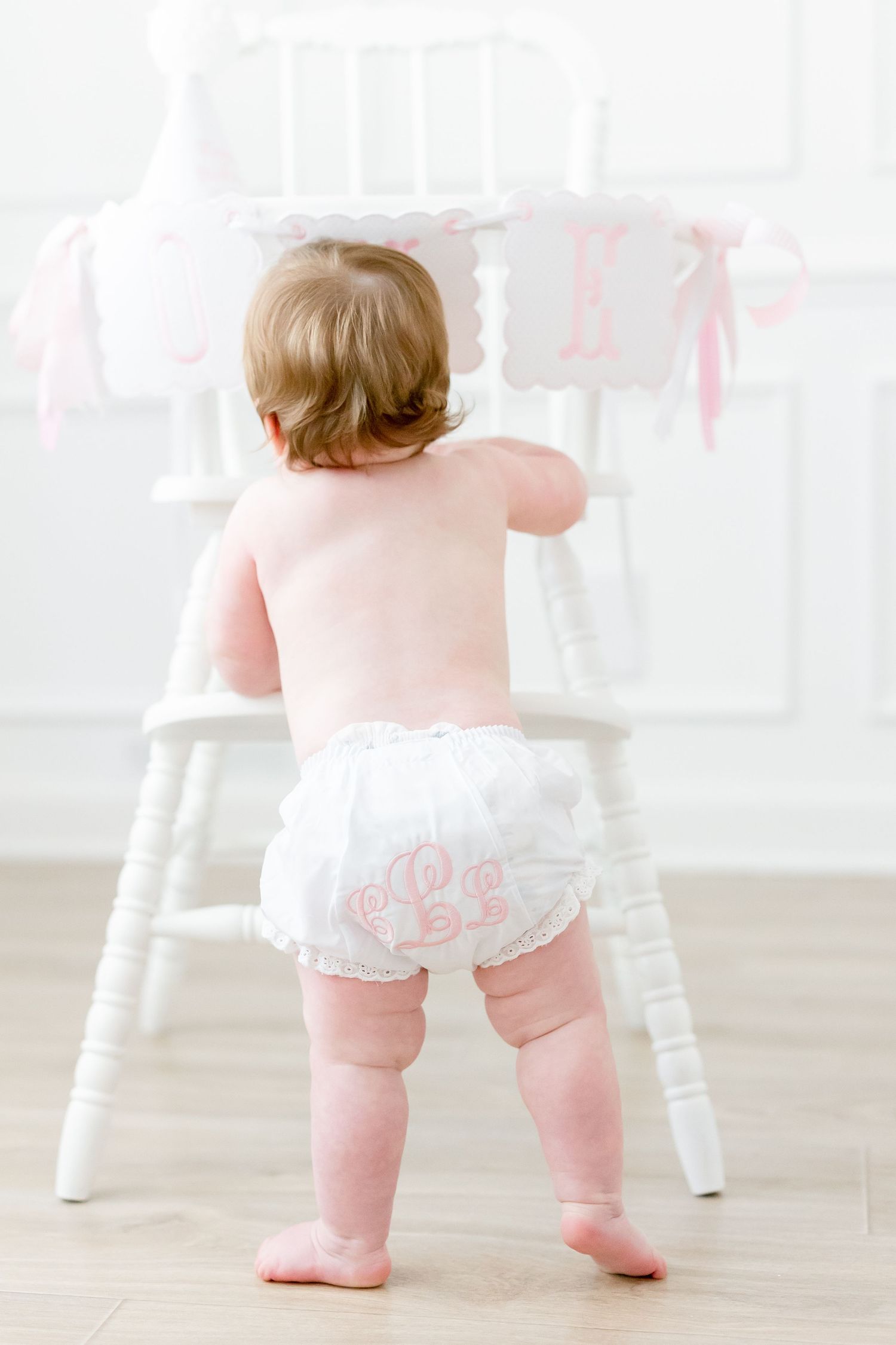 Baby girl in white monogrammed ruffle bloomers standing at a white chair with a pink birthday banner.