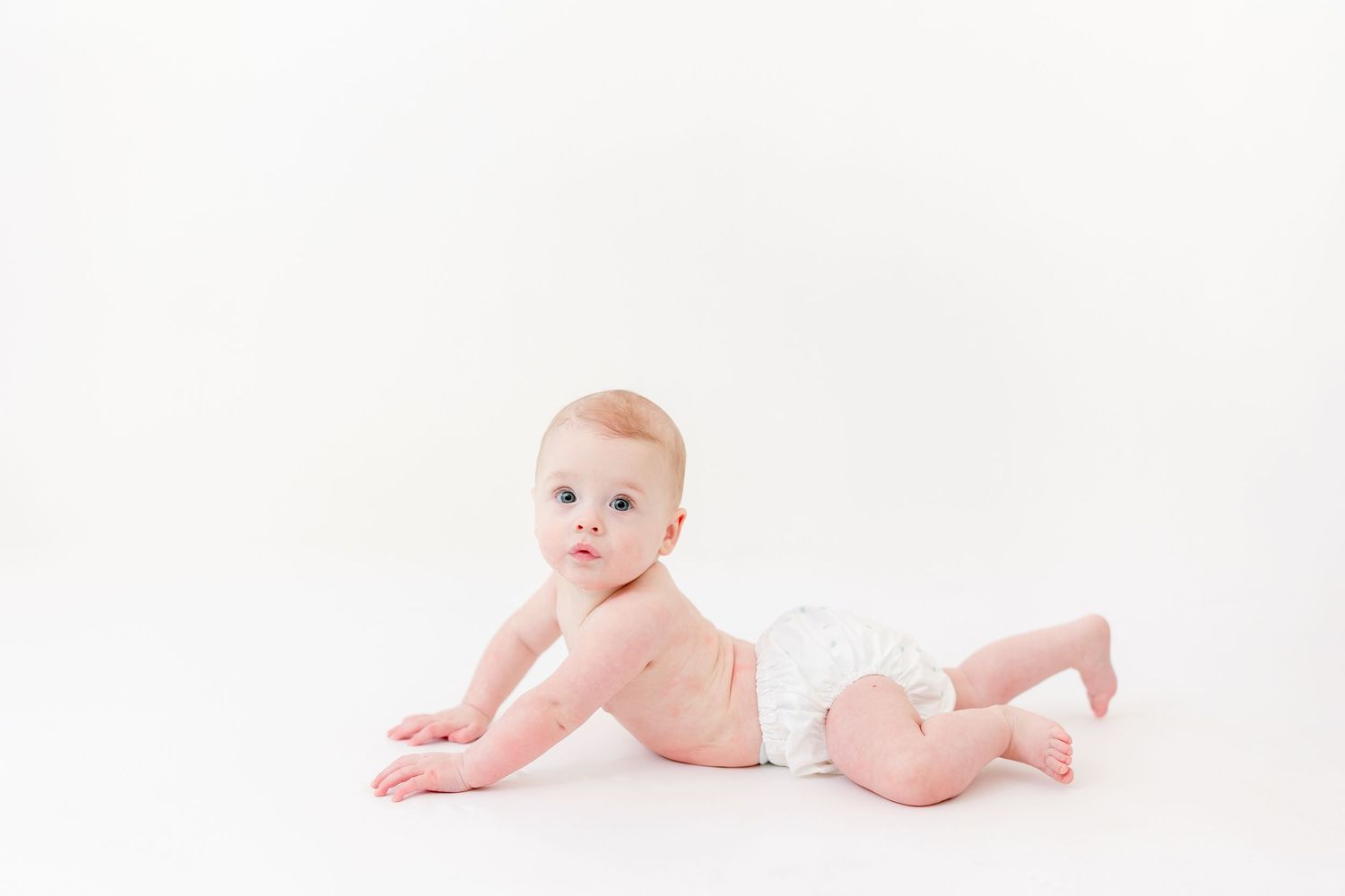 Smiling baby in a white diaper lying on their tummy and propped up on their arms against a clean white studio background.