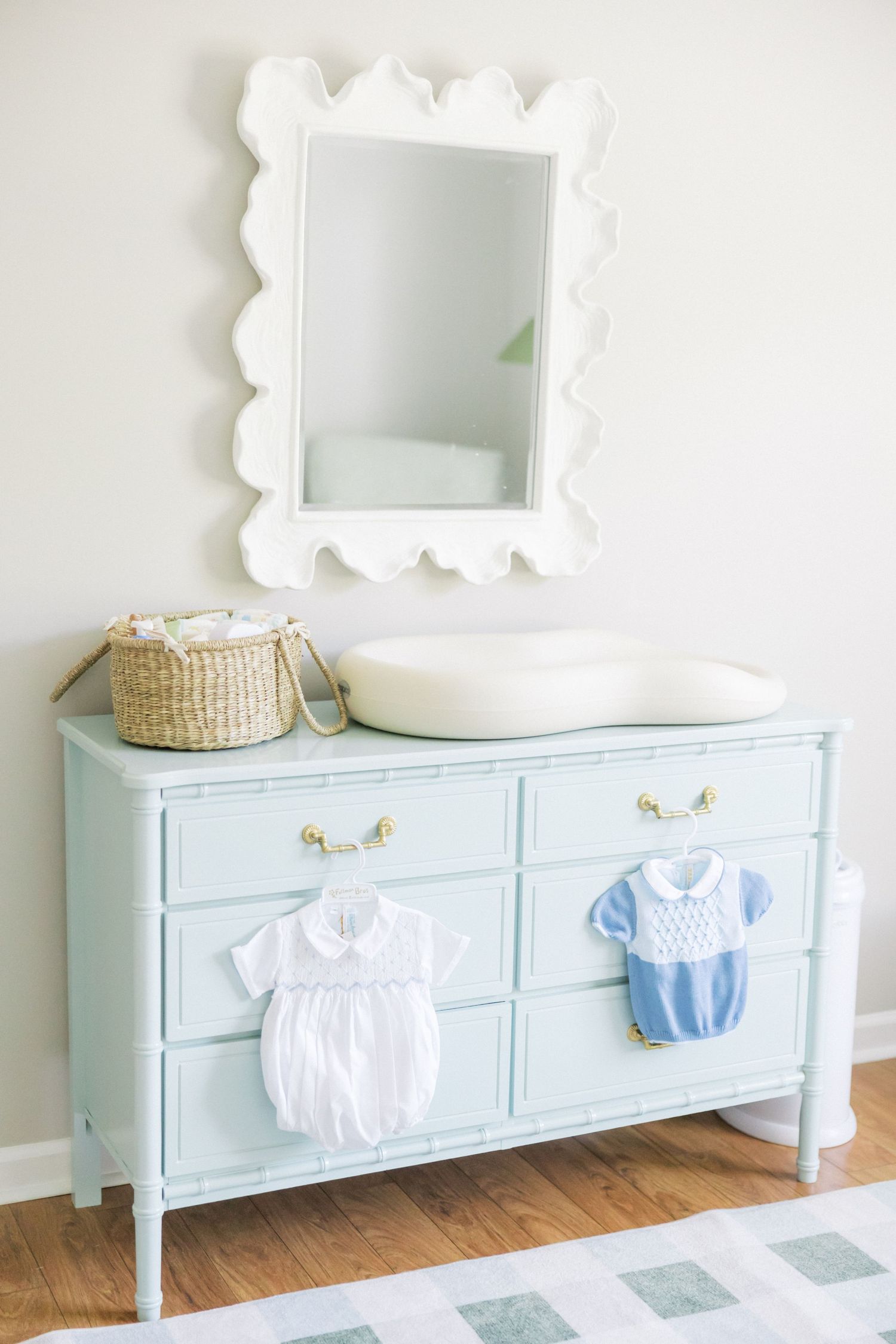 Light blue vintage dresser with decorative mirror displays baby rompers and basket in a minimalist nursery setting.