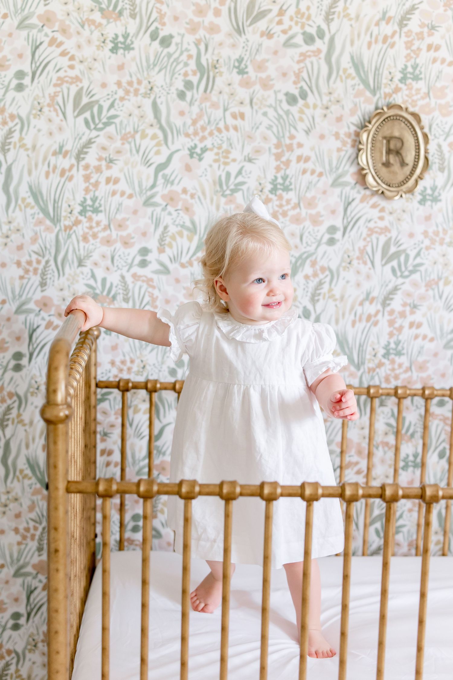 A small child in a white dress stands against a floral wallpaper background near a vintage wooden crib.