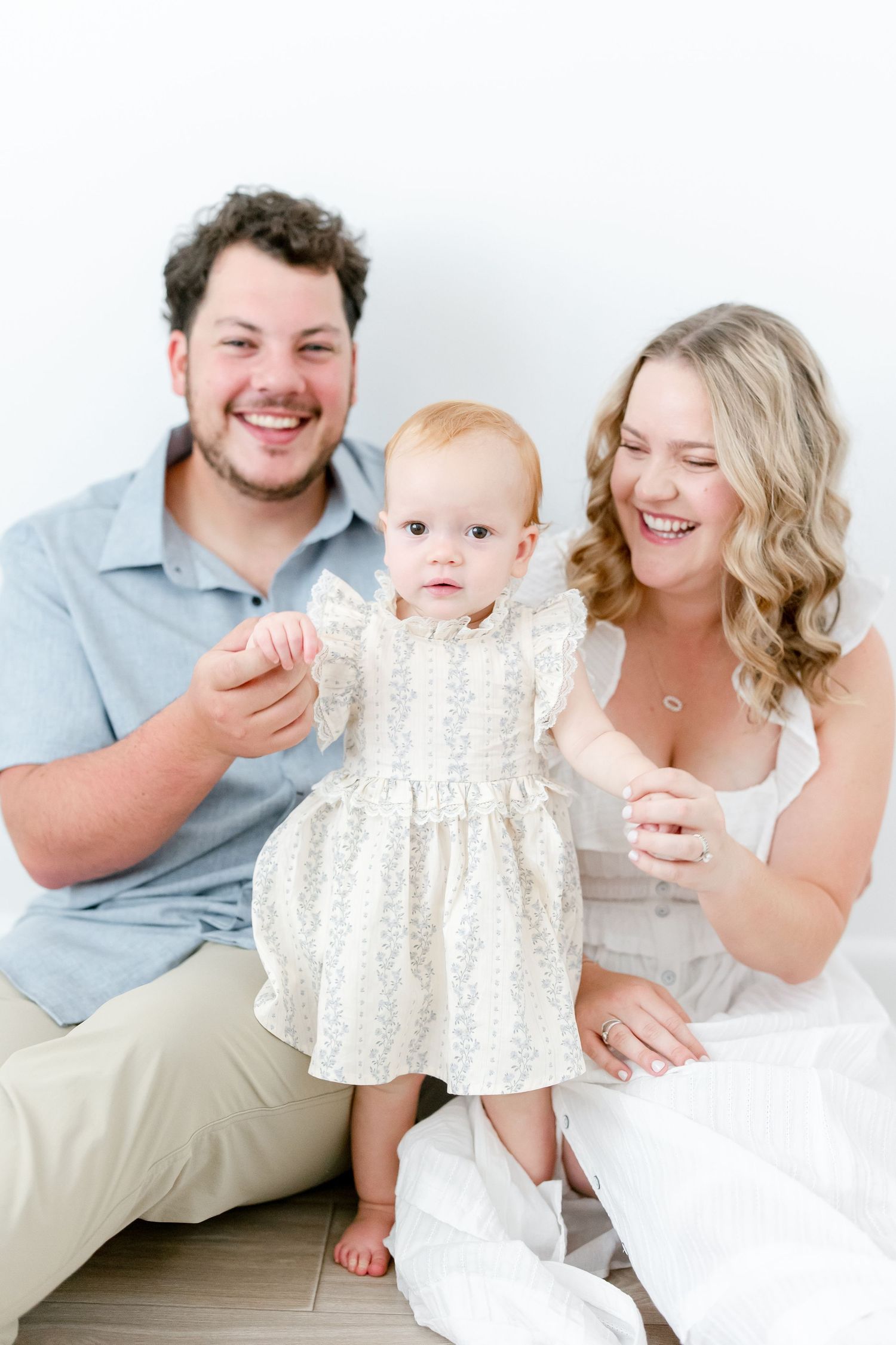 Happy family portrait with baby wearing white lace dress as parents hold them while sitting and laughing together.