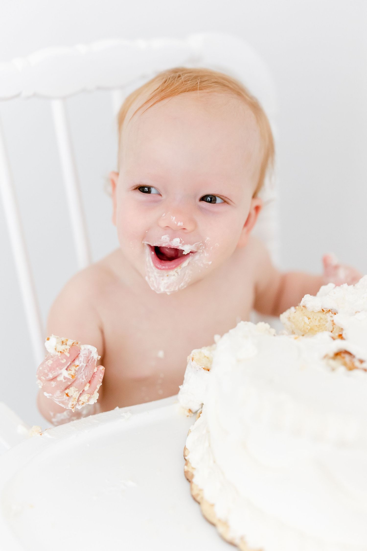 Happy baby in a white high chair enjoying their first birthday cake smash and making a wonderful mess.