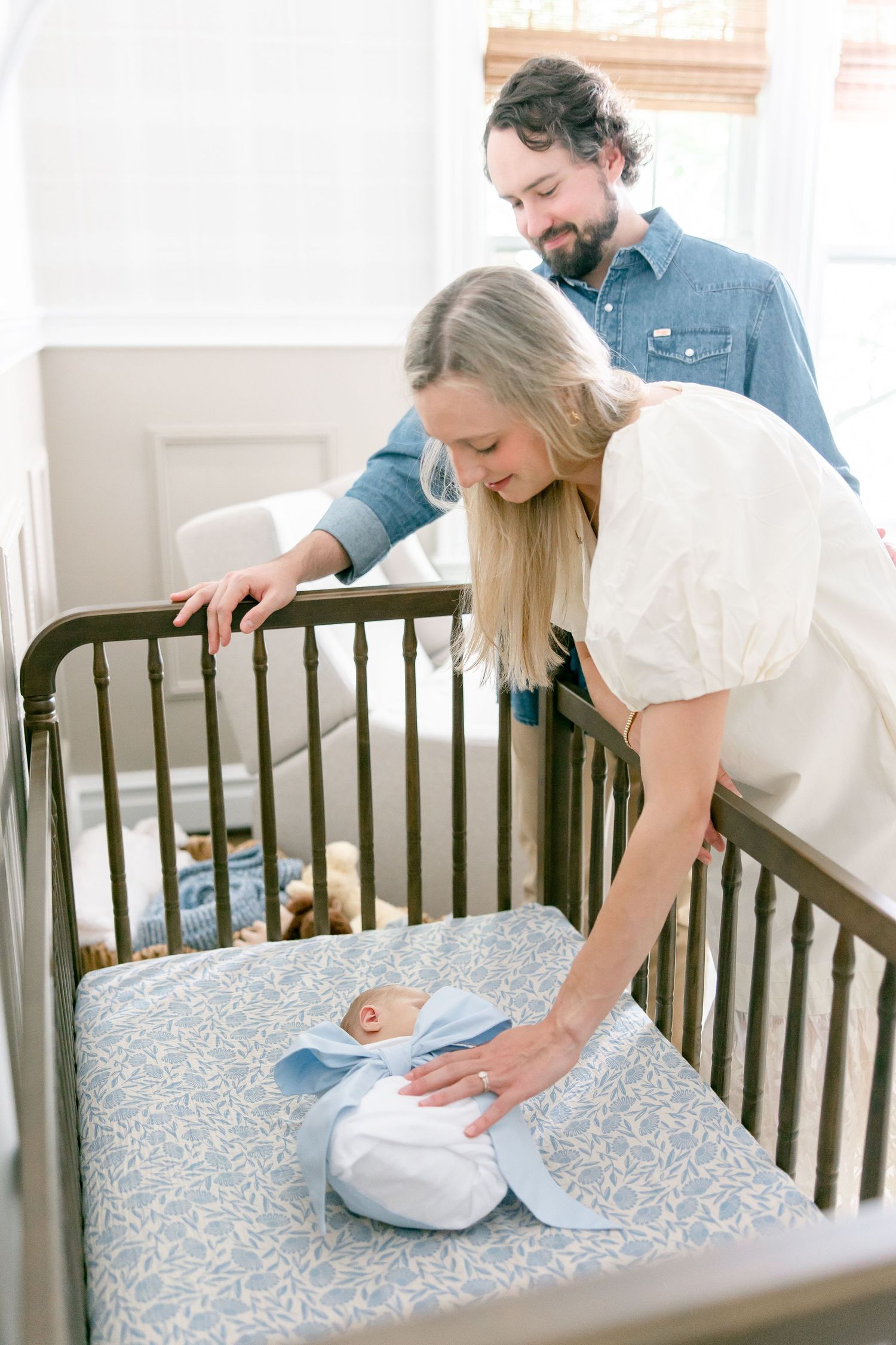 Parents lean over dark wood crib with floral bedding while tending to their newborn.