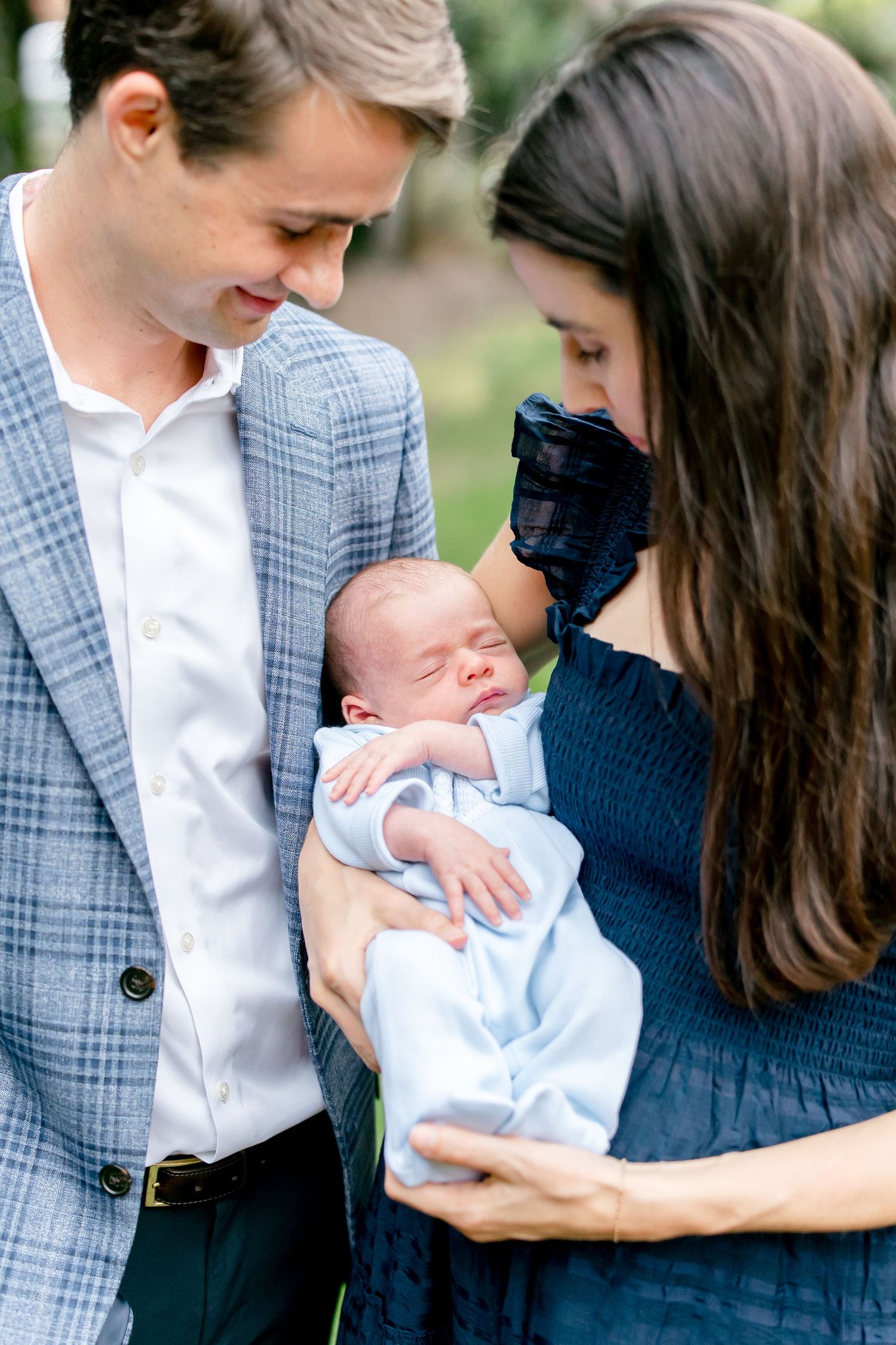 A couple holds and gazes at their newborn baby while wearing coordinating navy and gray outfits in an outdoor setting.