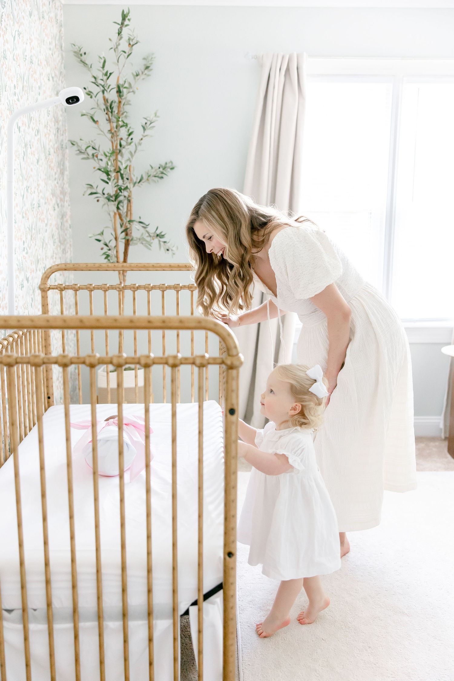 Parent gently interacts with baby in white nursery featuring natural rattan crib and potted bamboo plants near window.