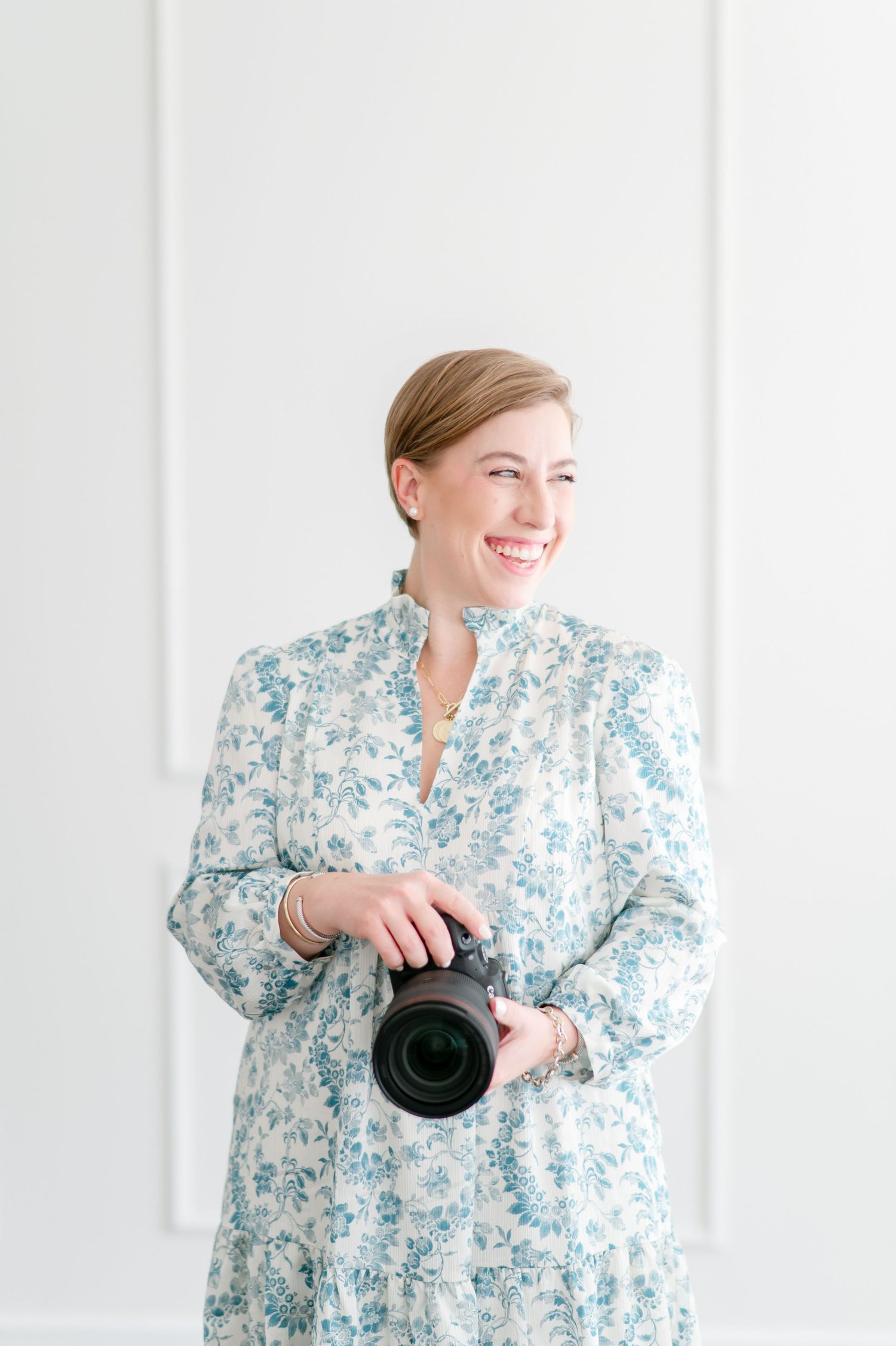 Professional photographer in a blue and white floral dress holding a camera while smiling against white wall.