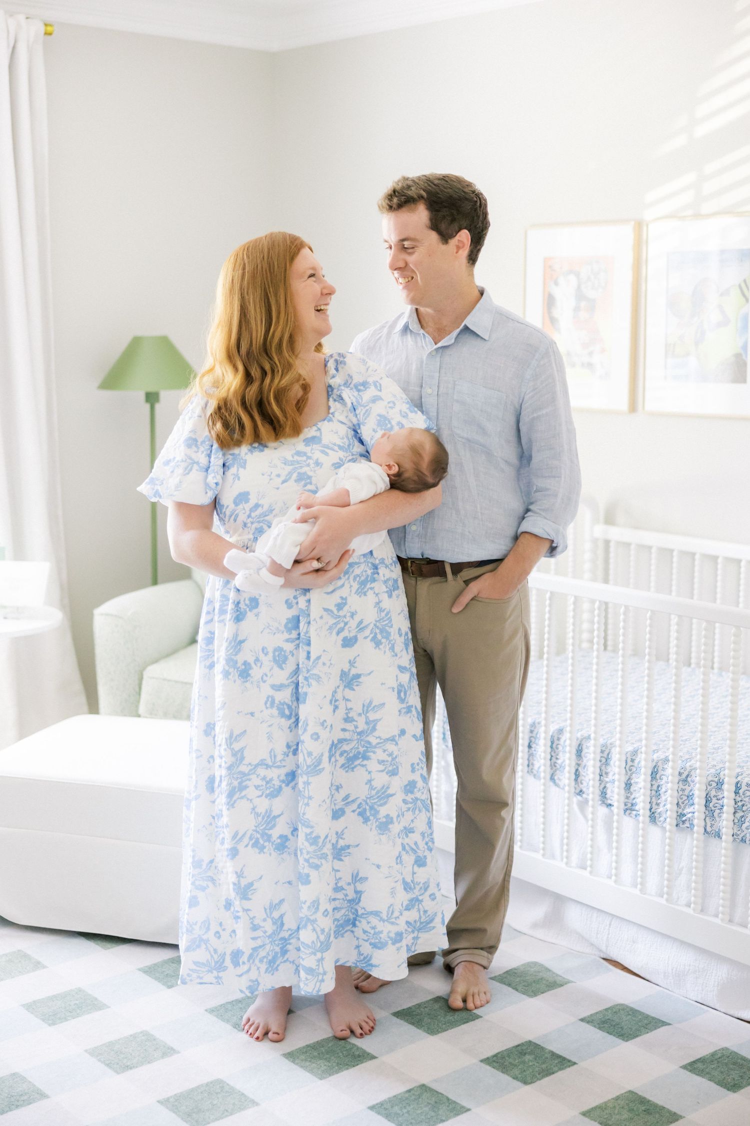 A couple stands in a bright nursery with a newborn baby, wearing blue and white while standing on decorative tile flooring.