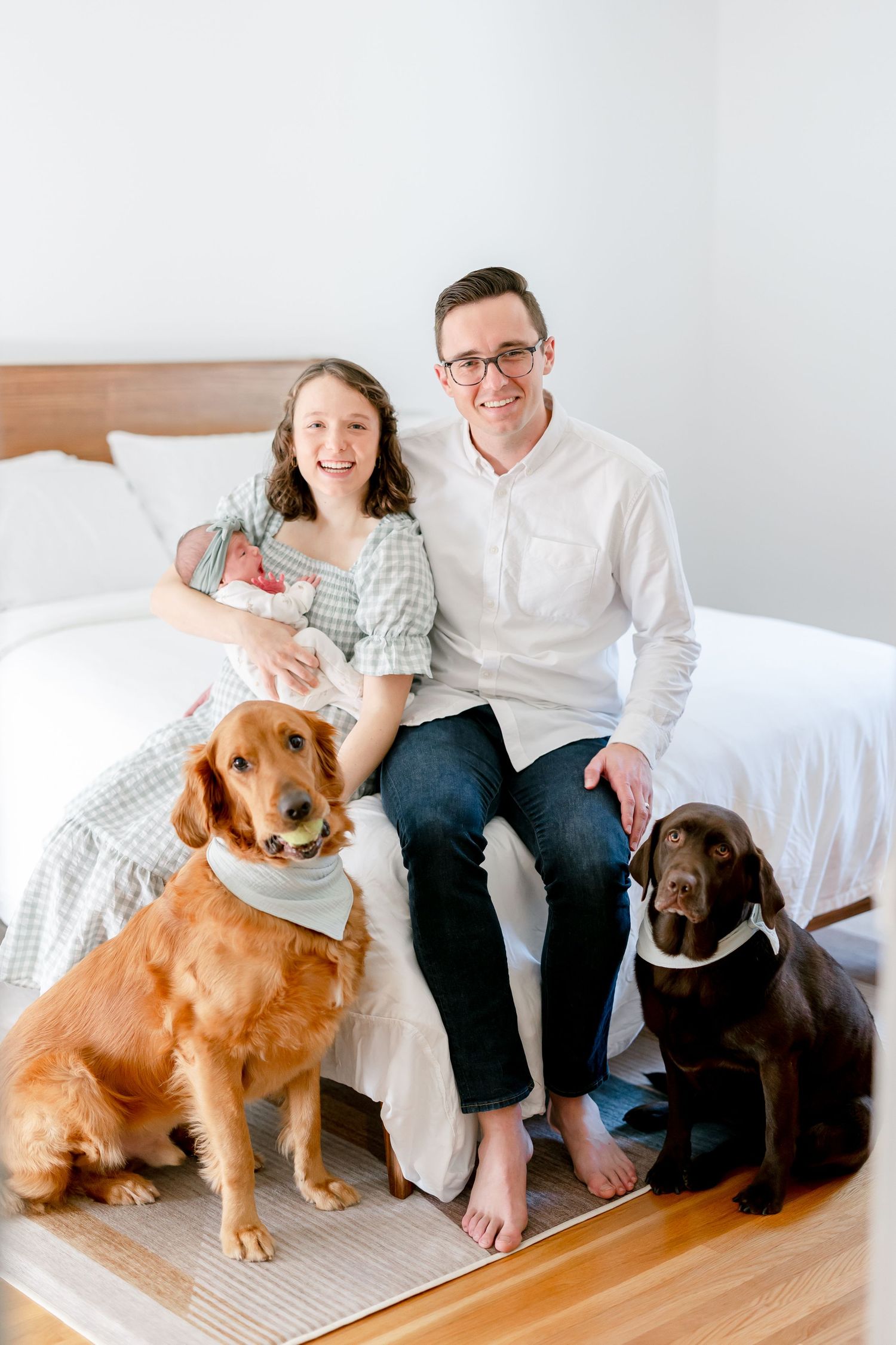 A family sits on a bed with their two dogs - a golden retriever and chocolate lab wearing white bandanas.